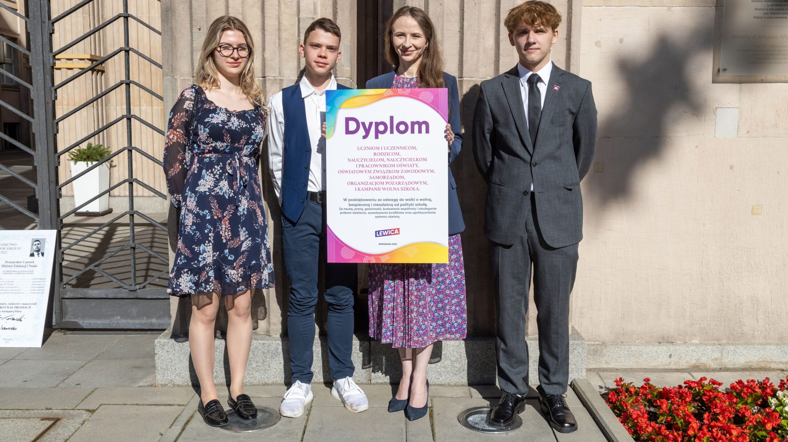 Cztery osoby stoją przed kamiennym budynkiem, trzymają duży kolorowy plakat z napisem "Dyplom".