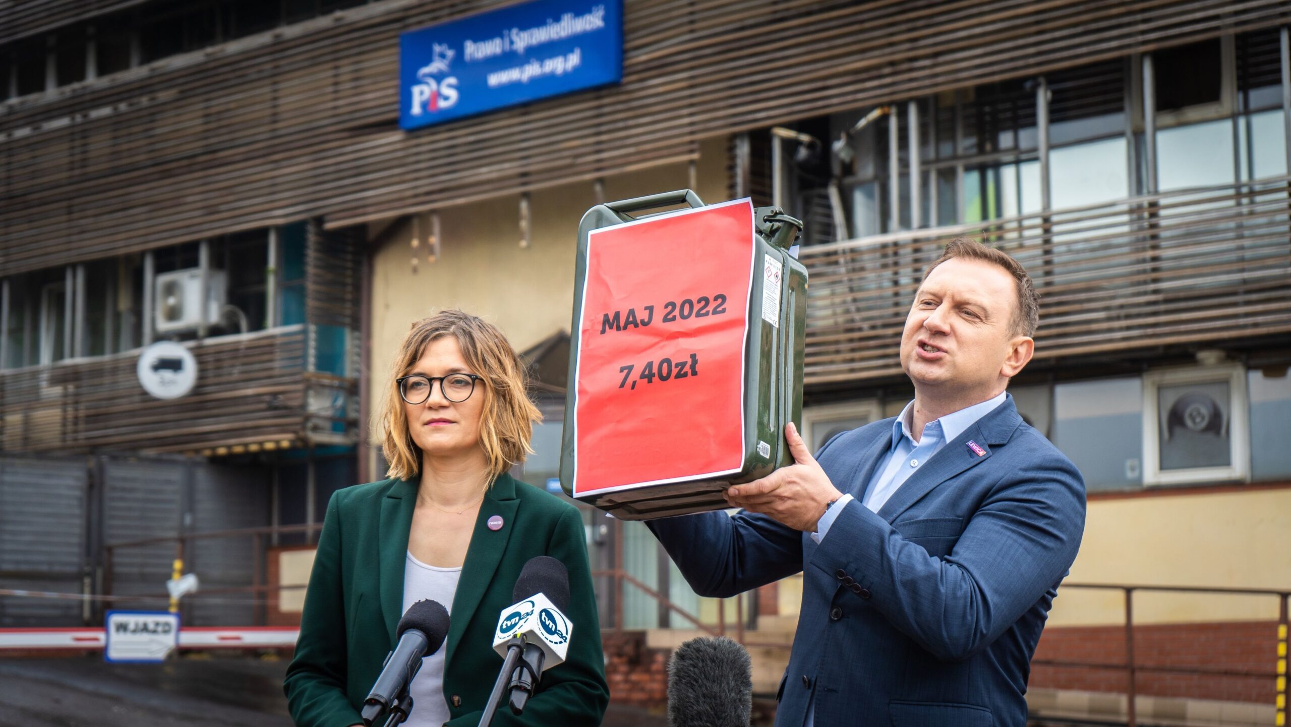 Na zdjęciu przedstawiono dwóch mówców, z których jeden trzyma kanister z czerwonym napisem, informującym o cenie oraz dacie, podczas konferencji prasowej przed budynkiem z logiem partii politycznej.