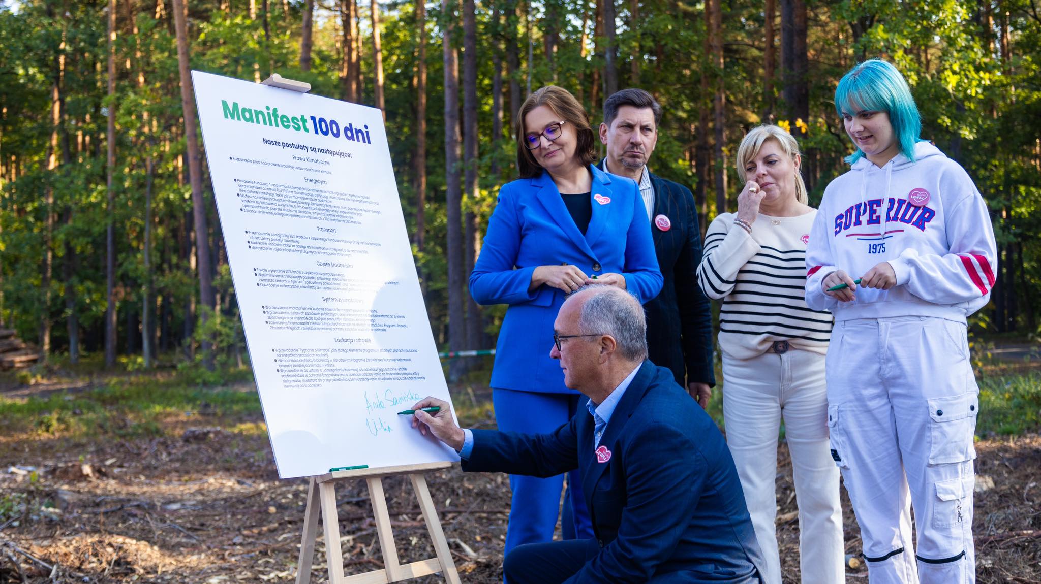 Grupa osób w lesie bierze udział w podpisywaniu manifestu „Manifest 100 dni dla polskiej przyrody i klimatu” przy dużej planszy ustawionej na sztaludze.