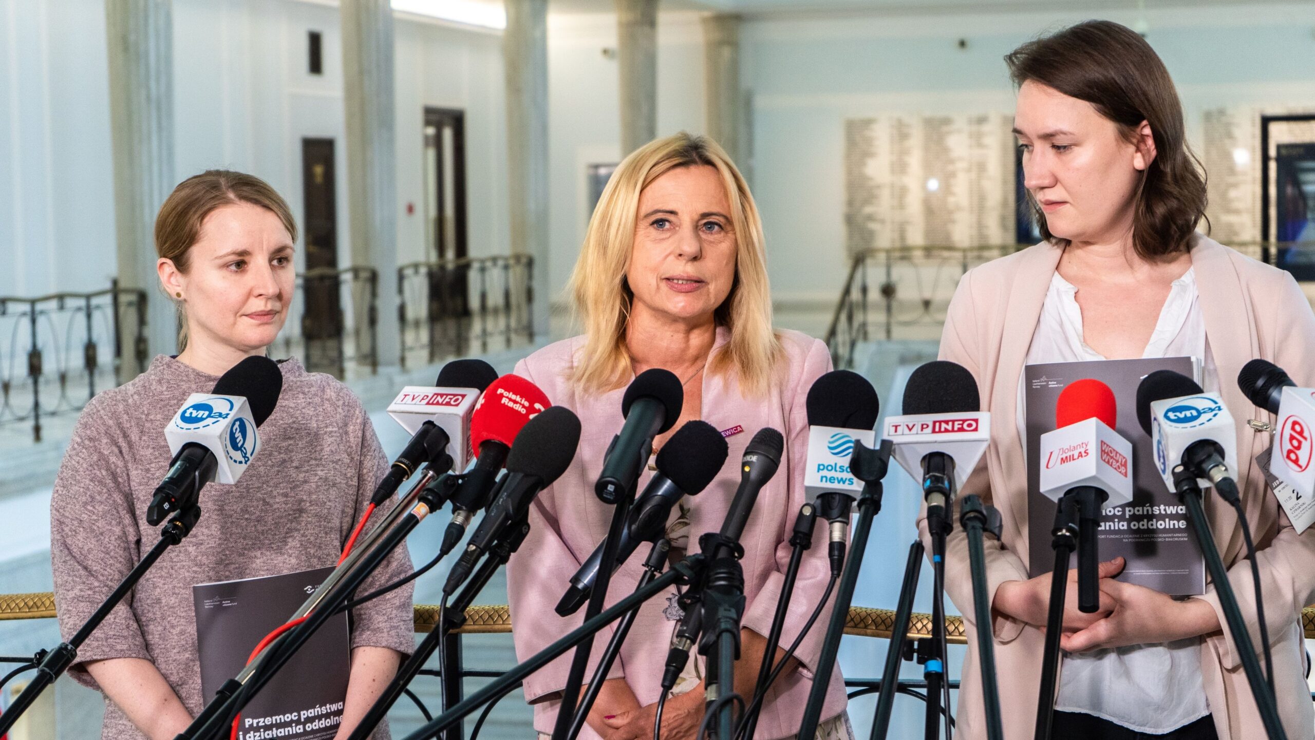 Na zdjęciu znajdują się trzy kobiety w trakcie konferencji prasowej, stojące przed mikrofonami z różnych mediów, w tle widać przestronną salę.