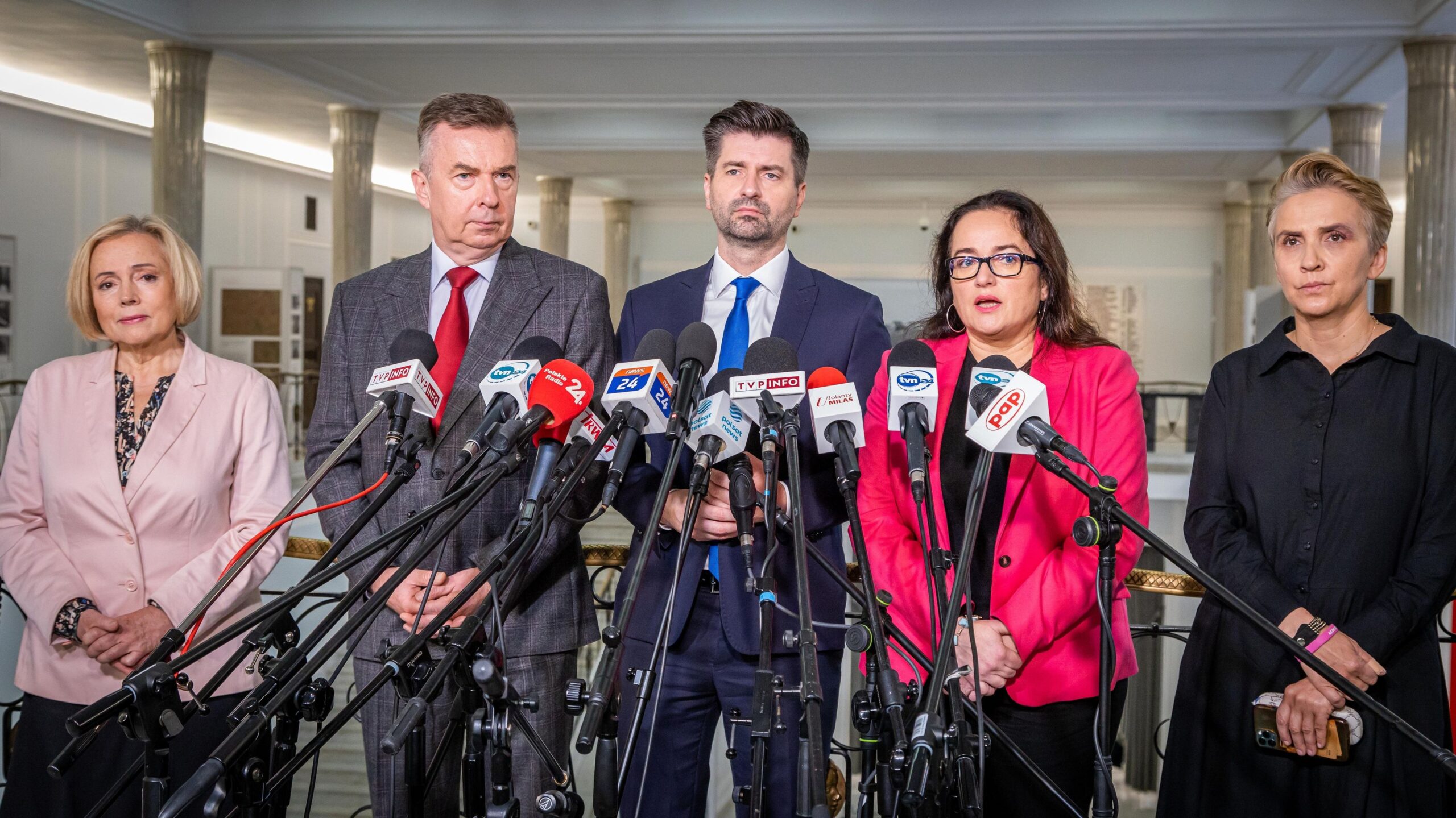 Na zdjęciu znajduje się pięć osób stojących przed mikrofonami, w trakcie konferencji prasowej w jasnym, eleganckim wnętrzu.