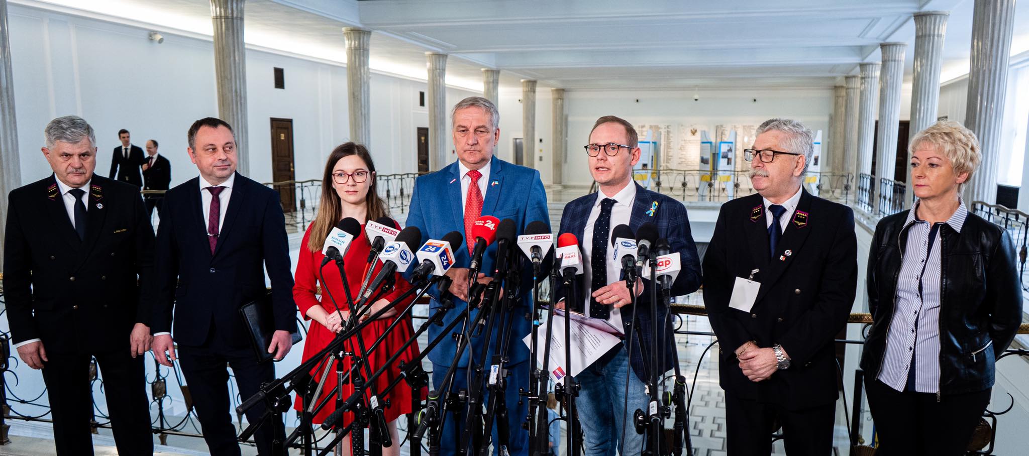 Na zdjęciu znajduje się grupa ośmiu osób stojących razem w pomieszczeniu z mikrofonami, podczas konferencji prasowej.