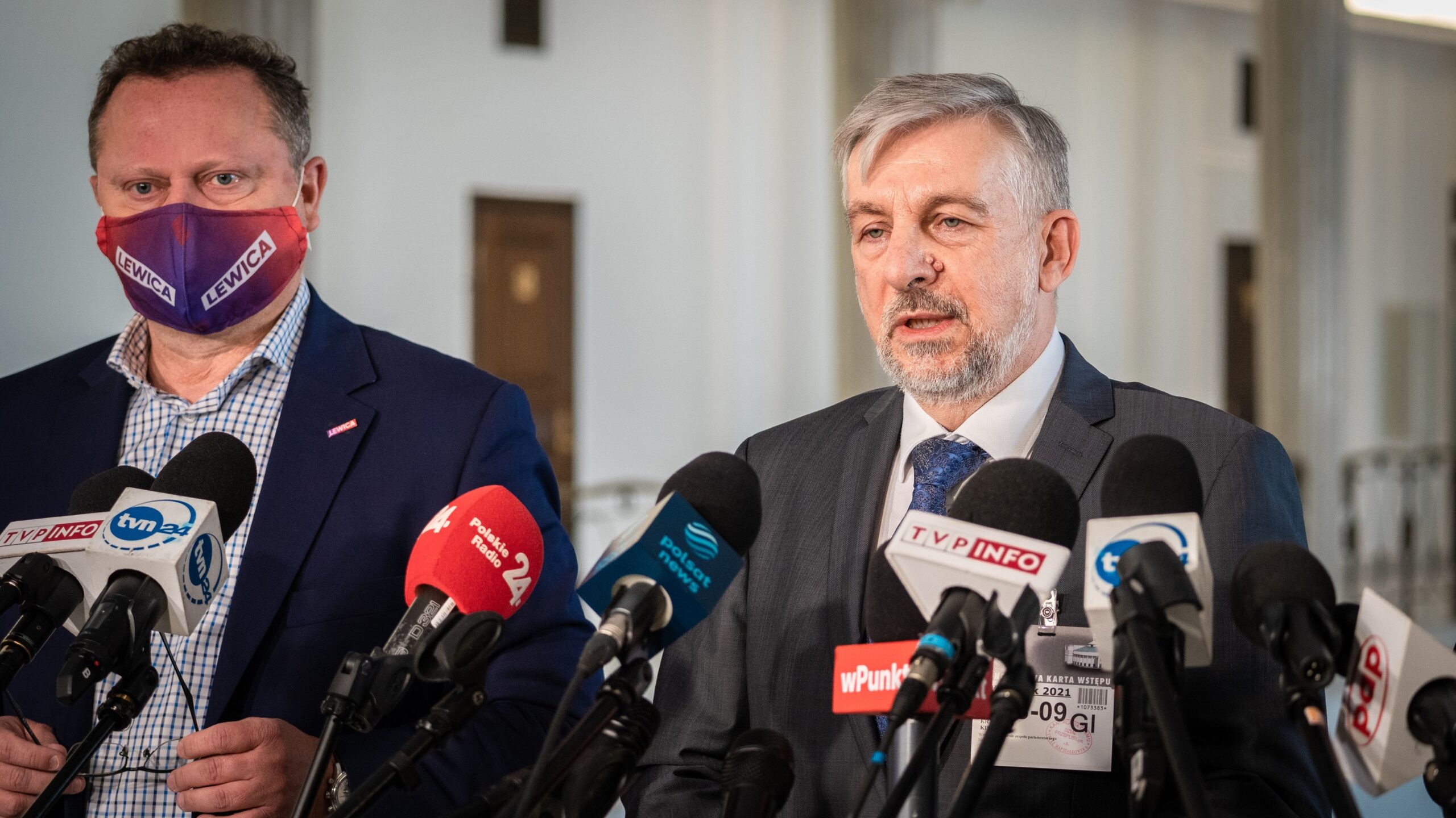 Dwóch mężczyzn w garniturach podczas konferencji prasowej, stojących za licznymi mikrofonami; jeden z nich ma maseczkę z logo Lewica.