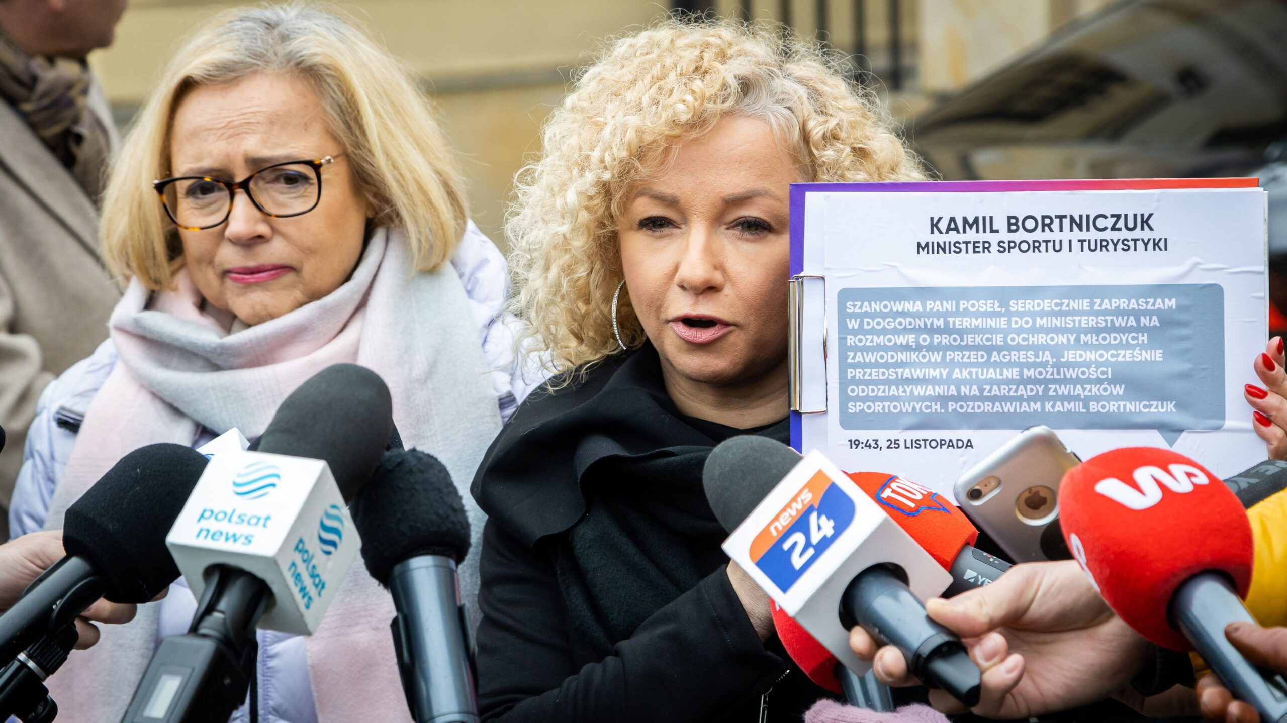 Na zdjęciu dwie kobiety rozmawiają z mediami podczas konferencji, trzymając dokument z zaproszeniem do rozmowy na temat ochrony młodych zawodników w sporcie.