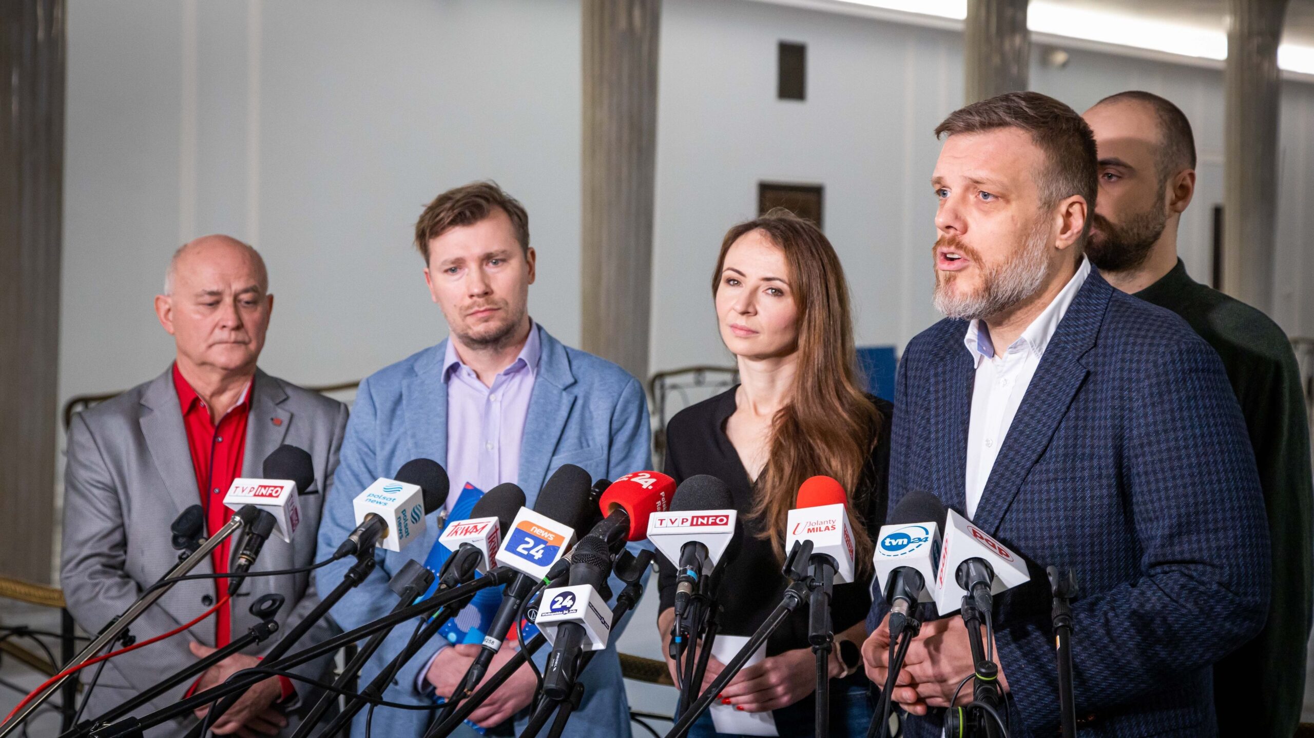 Konferencja prasowa z udziałem kilku osób stojących za mikrofonami, z których część jest skupiona na wystąpieniu mężczyzny z brodą.