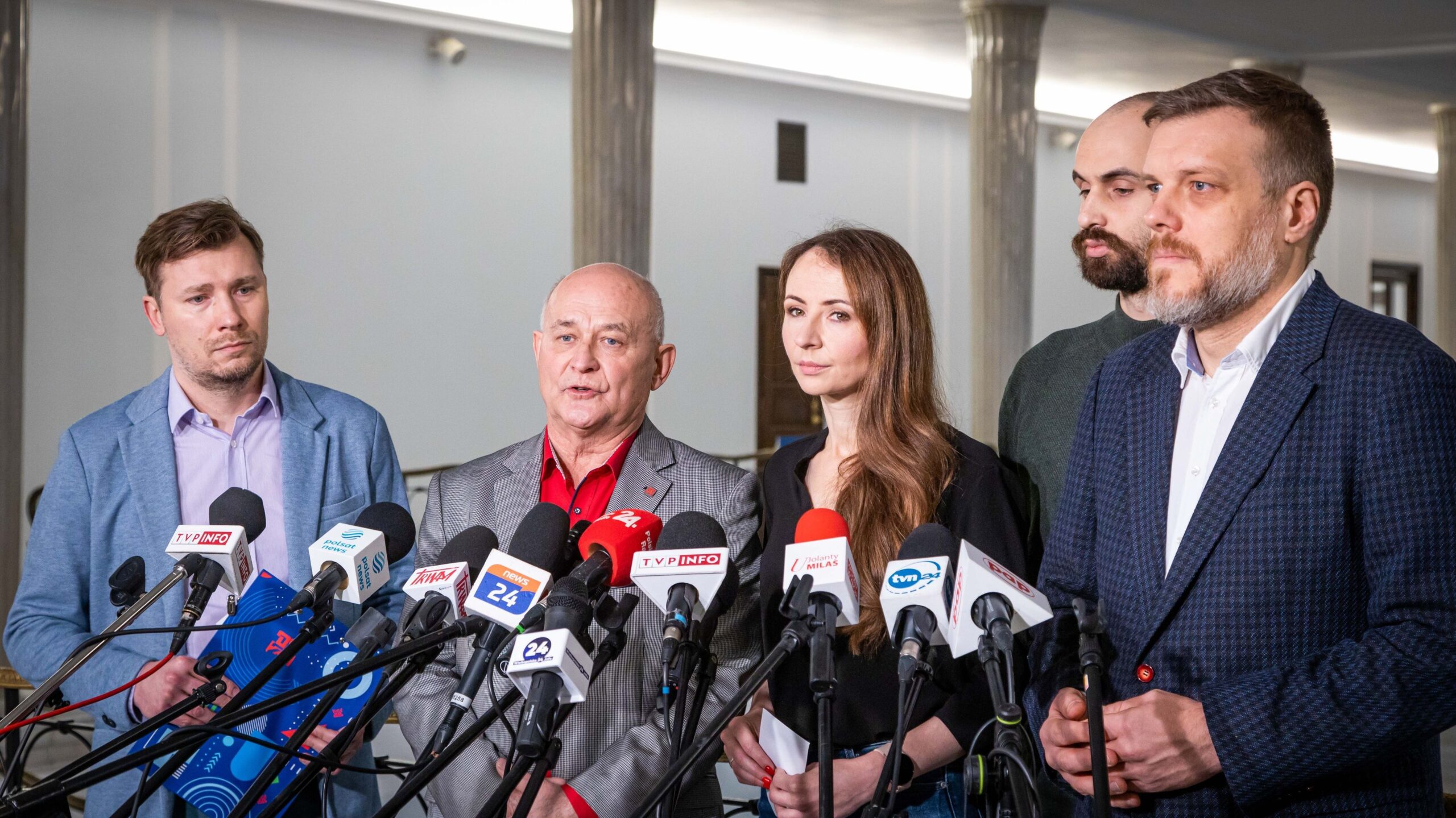 Grupa pięciu osób w biznesowych ubraniach stoi na konferencji prasowej otoczona licznymi mikrofonami z logo mediów, omawiając temat pracy platformowej.