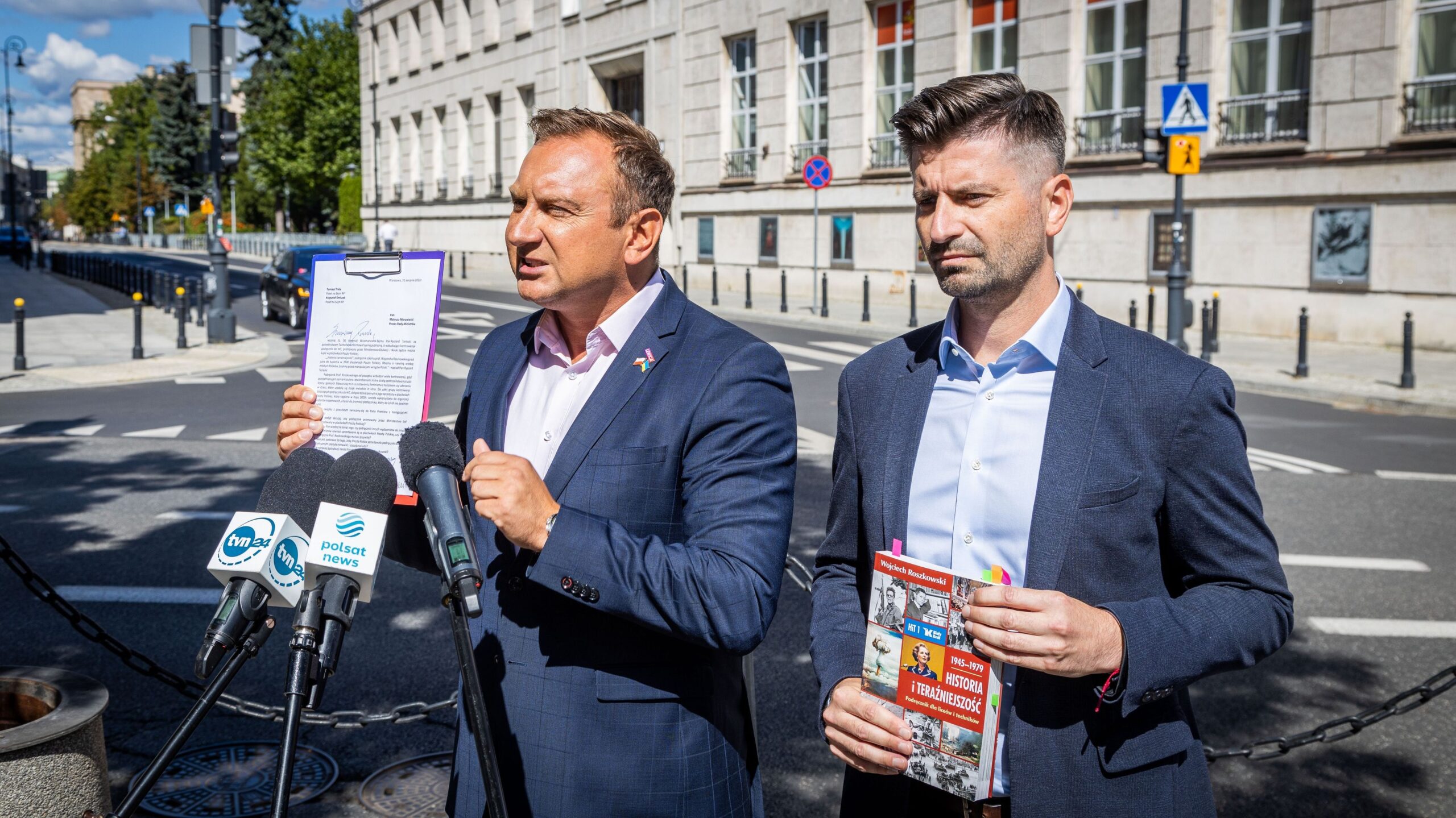 Na zdjęciu dwóch mężczyzn prowadzi konferencję prasową na ulicy, trzymając dokumenty oraz broszurę.