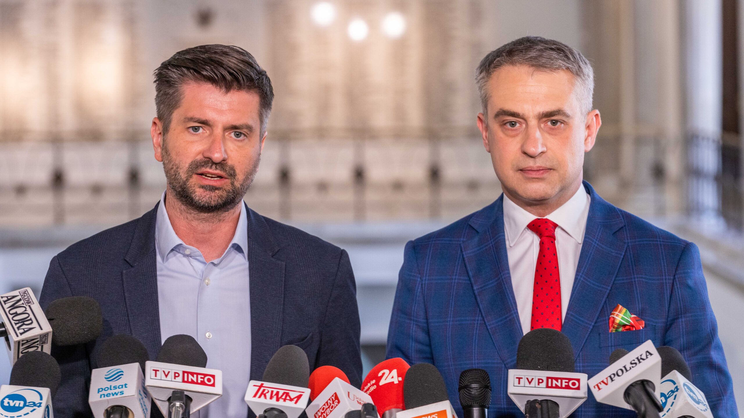 Dwóch mężczyzn w garniturach stoi przed zestawem mikrofonów podczas konferencji prasowej.