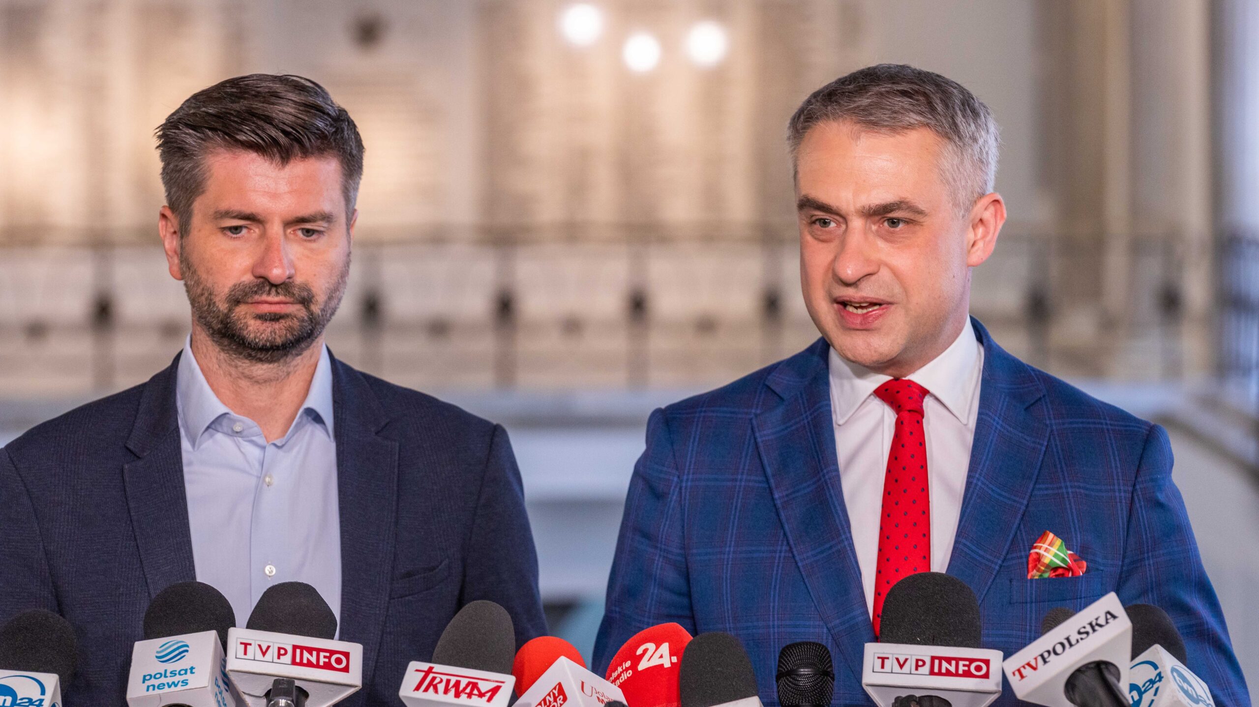 Dwóch mężczyzn w garniturach udziela konferencji prasowej, stojąc przed szeregiem mikrofonów.