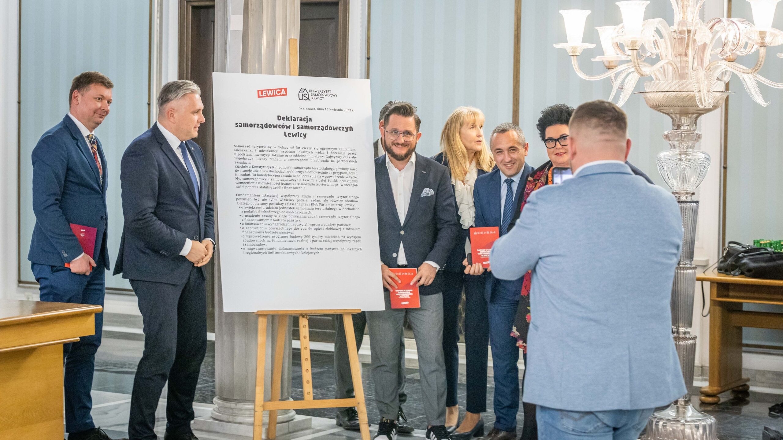 Grupa osób w garniturach stoi przy plakacie z deklaracją samorządowców Lewicy podczas konferencji, pod hasłem Silny samorząd to silna Polska.