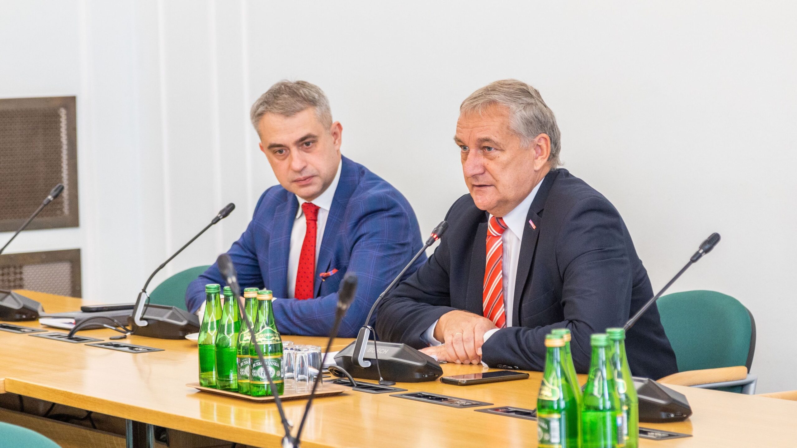 Na zdjęciu widać dwóch mężczyzn siedzących przy stole, z butelkami wody mineralnej w tle, podczas spotkania lub konsultacji.