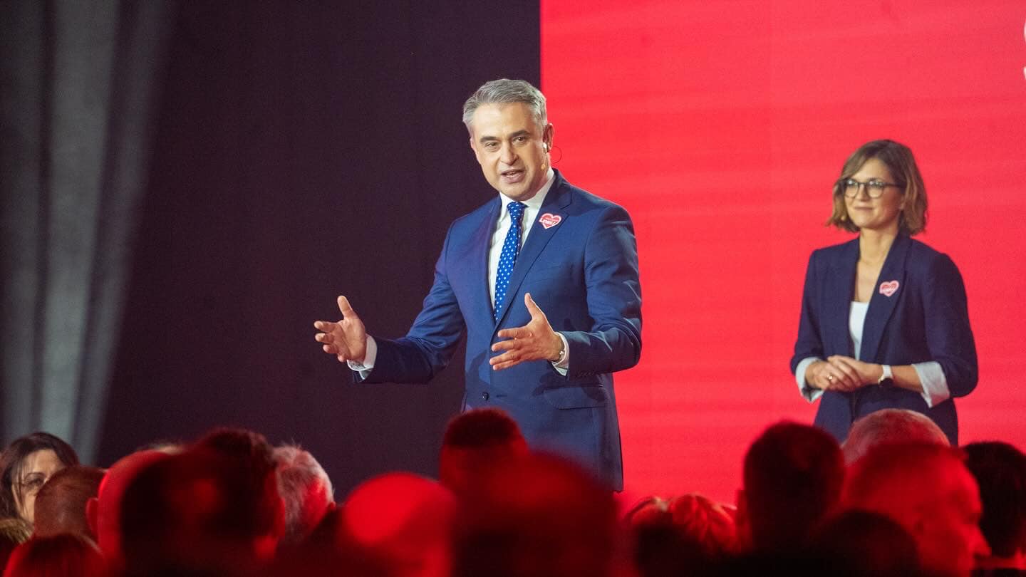 Na scenie podczas konwencji samorządowej Lewicy mężczyzna w granatowym garniturze wygłasza przemówienie, a obok niego stoi kobieta w dopasowanym garniturze na tle czerwonej sceny, w otoczeniu zgromadzonej publiczności.