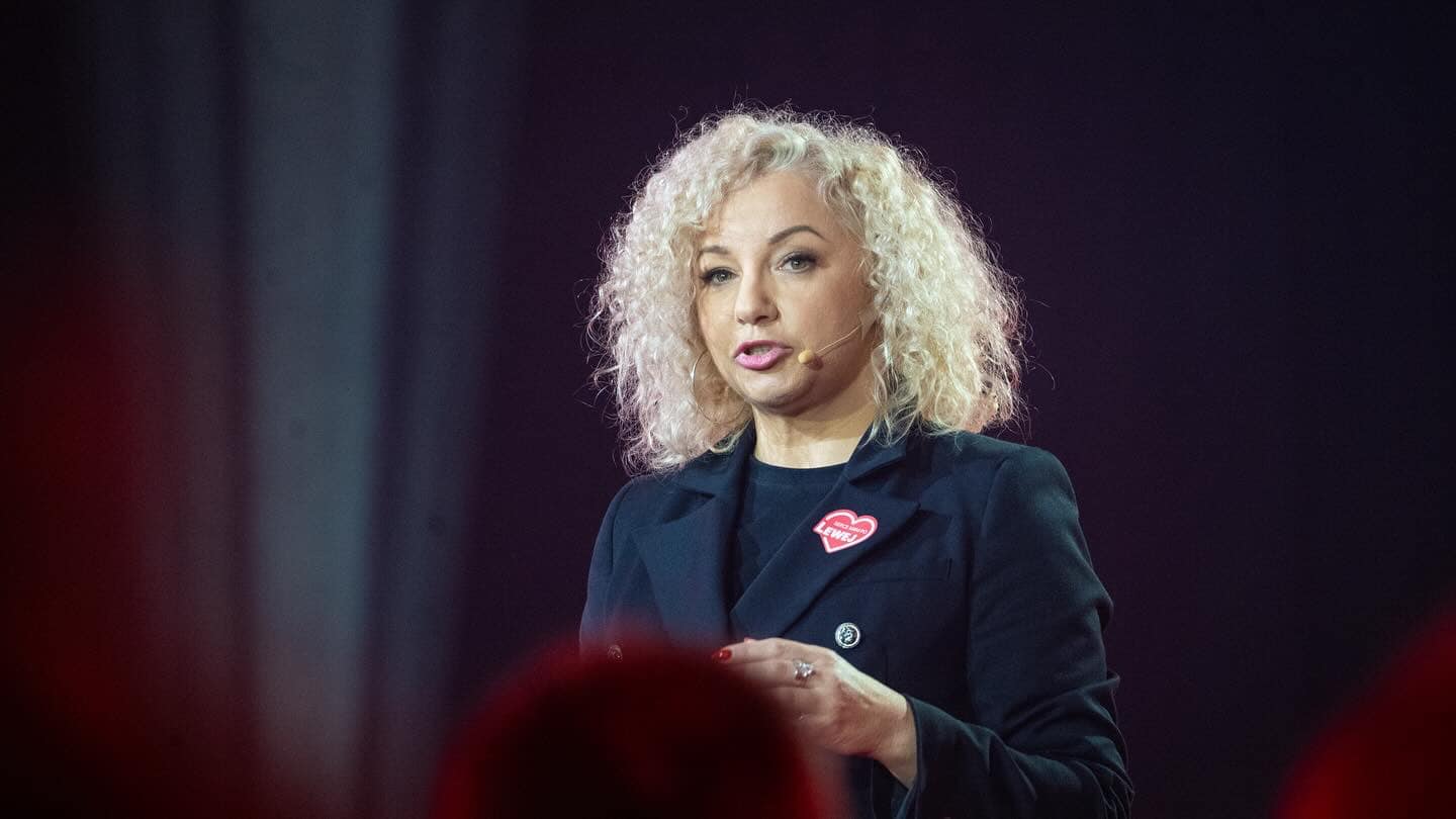 Kobieta z blond kręconymi włosami w czarnym żakiecie przemawia na scenie z mikrofonem, na klapie ma różowe serduszko Lewica podczas konwencji samorządowej Lewicy.