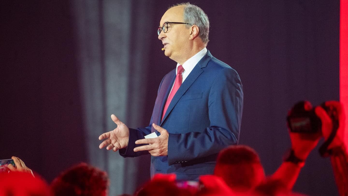 Mężczyzna w garniturze z czerwonym krawatem przemawia na scenie podczas konwencji samorządowej Lewicy.