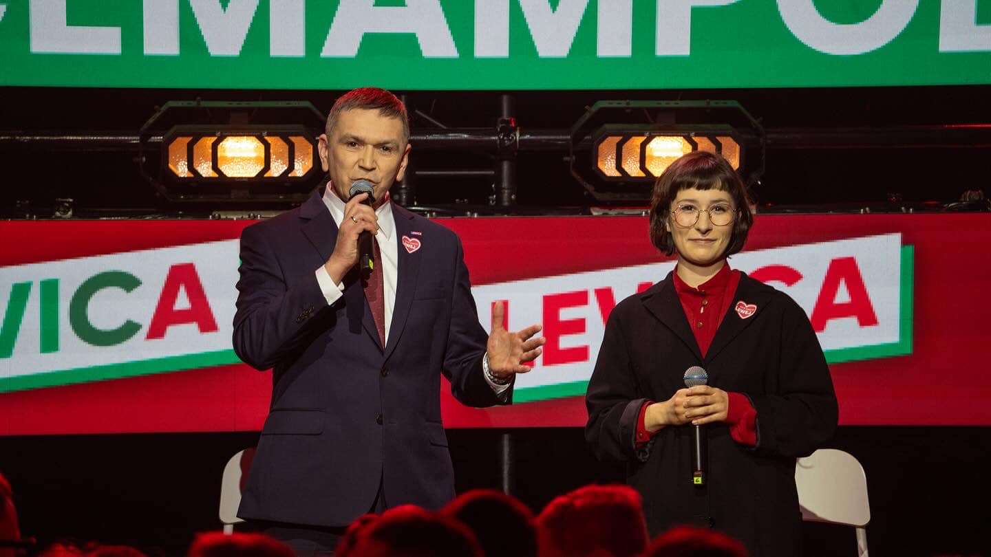 Dwóch prelegentów na scenie podczas konwencji samorządowej Lewicy, mężczyzna w garniturze przemawia do publiczności, kobieta obok trzyma mikrofon; w tle baner z napisem Lewica.
