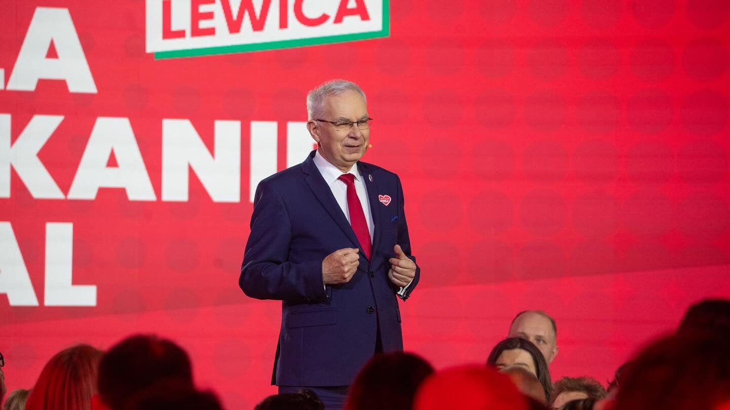 Mężczyzna w granatowym garniturze i czerwonym krawacie stoi na scenie podczas konwencji Lewicy, przed dużym czerwonym tłem z napisem LEWICA i zgromadzonymi widownią.