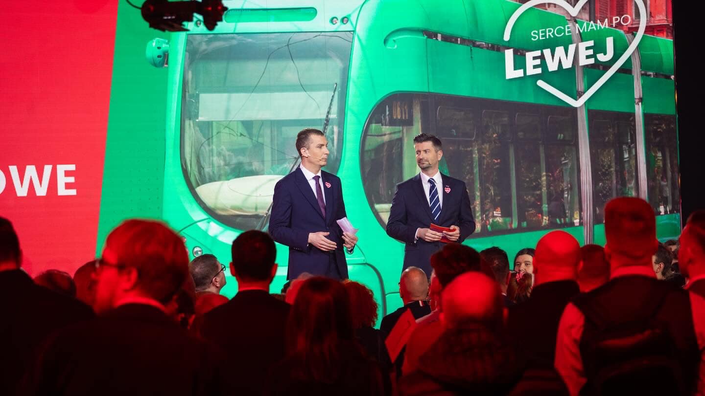 Dwóch mężczyzn w garniturach prowadzi przemówienie na scenie podczas konwencji Lewicy, przed dużym zielonym tłem z wizerunkiem tramwaju i logo partii.