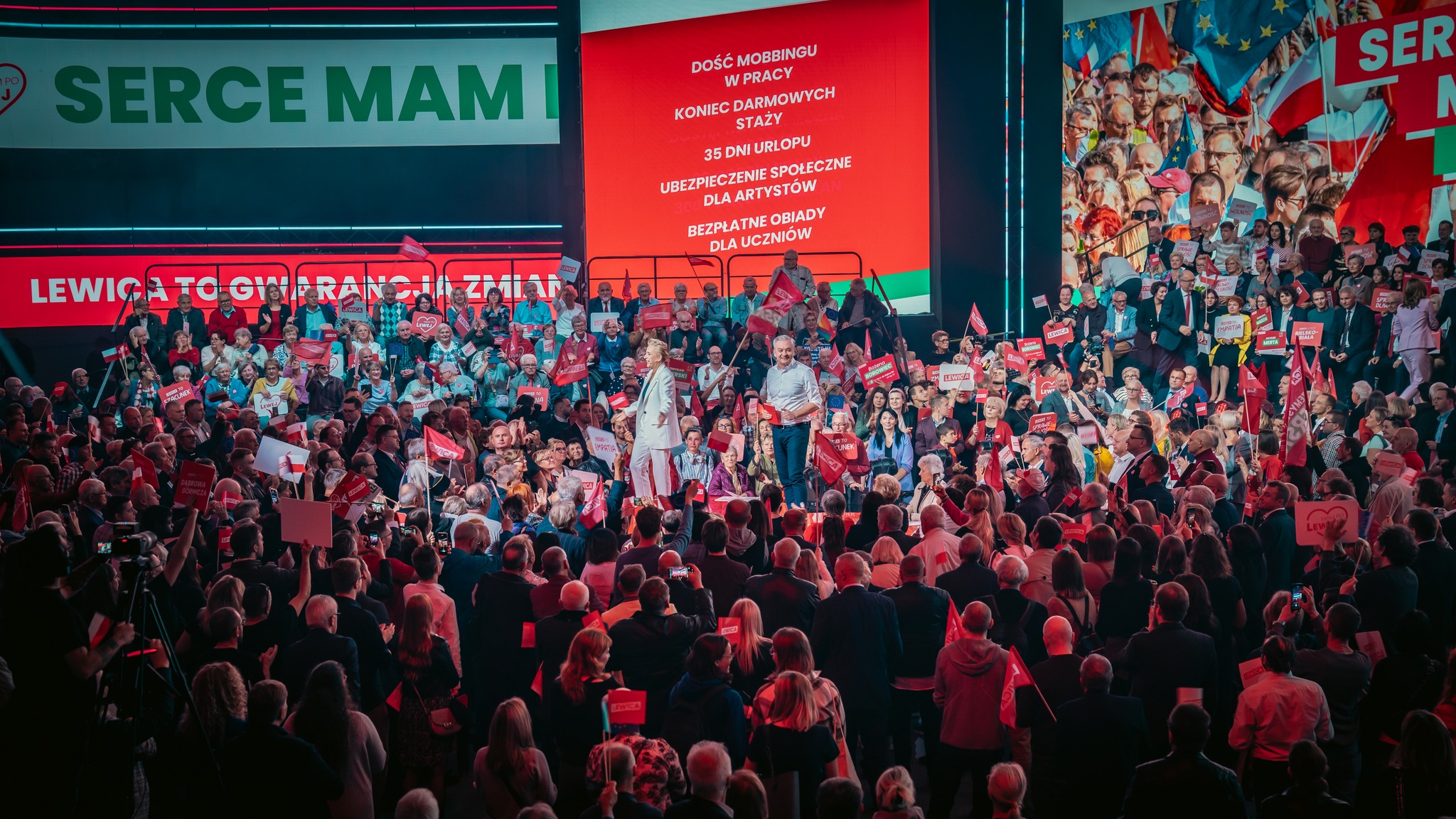 Ogromna publiczność na konwencji Lewicy w Będzinie, z czerwonymi flagami, przed sceną z dwoma mówcami i dużym czerwonym ekranem w tle.