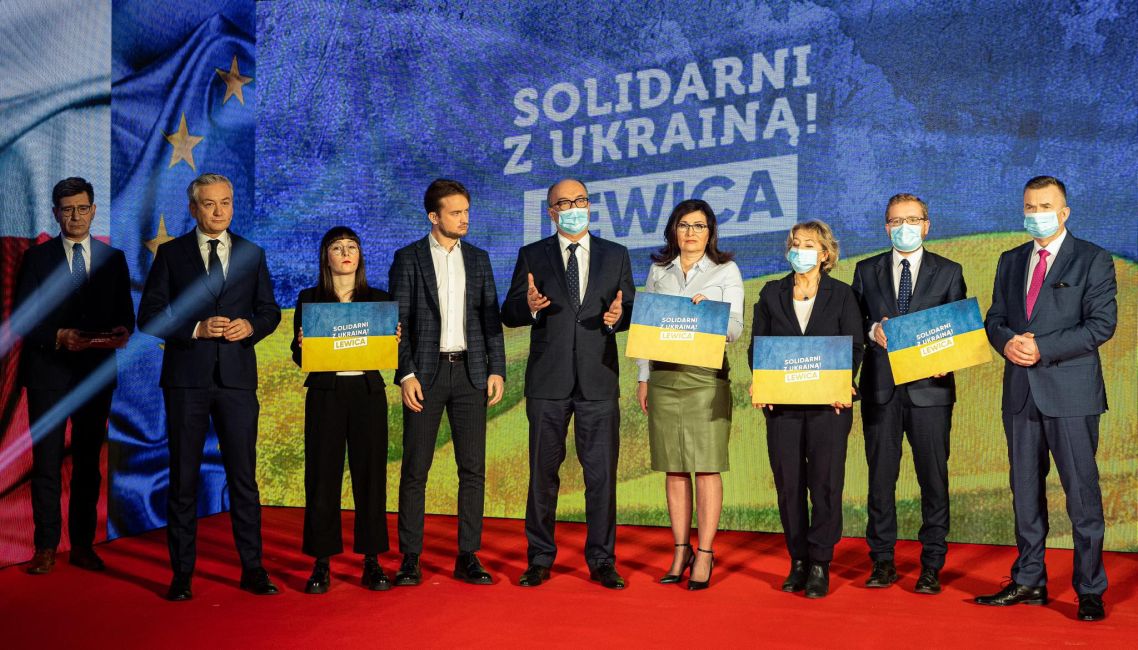 Na zdjęciu przedstawieni są członkowie partii Lewica w trakcie konferencji, trzymający transparenty z hasłem „Solidarni z Ukrainą!” na tle flagi polskiej i europejskiej.