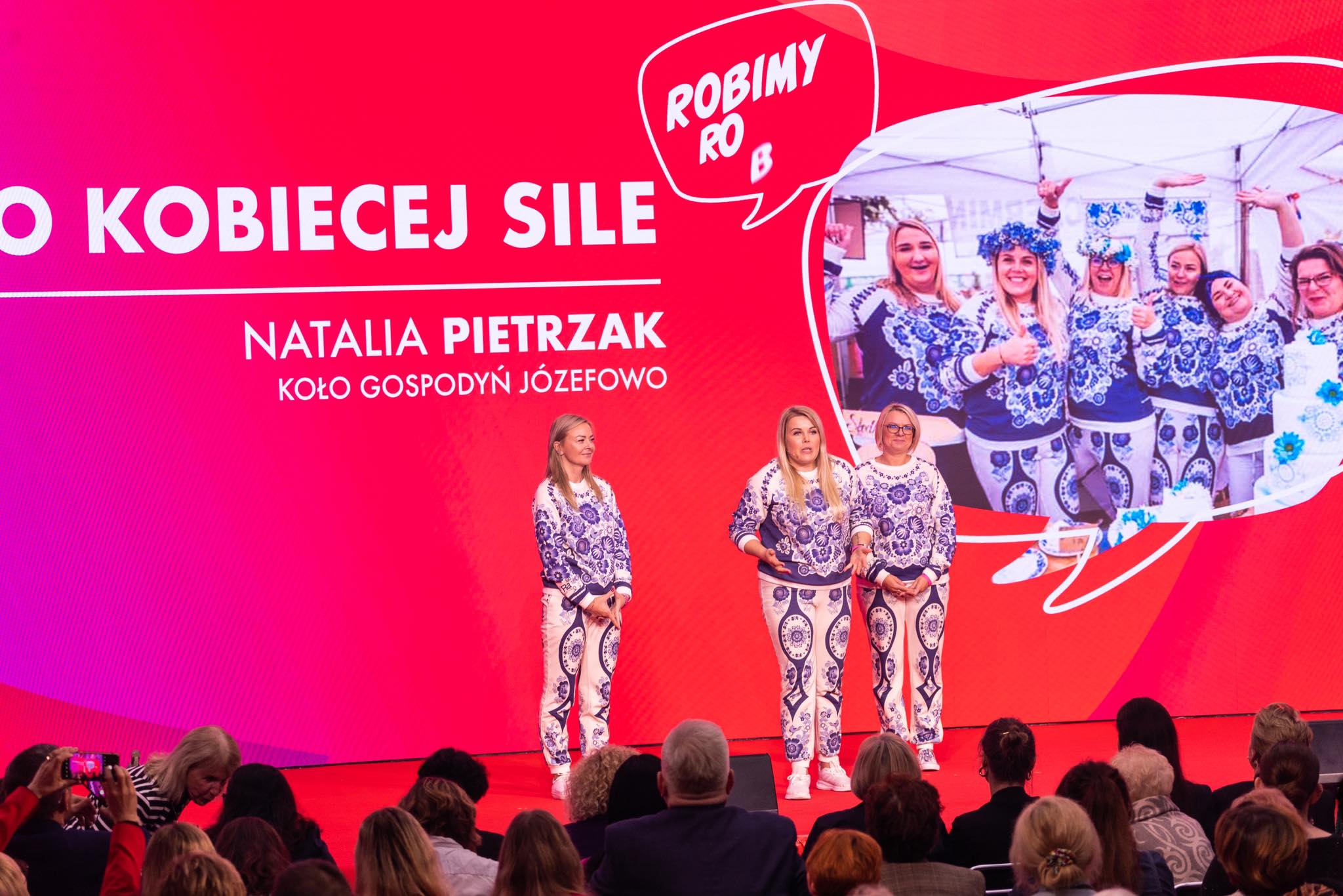 Na zdjęciu trzy kobiety w autentycznych strojach z ludowym wzorem występują na scenie podczas wydarzenia poświęconego sile kobiet, z czerwonym tłem i interaktywną prezentacją.