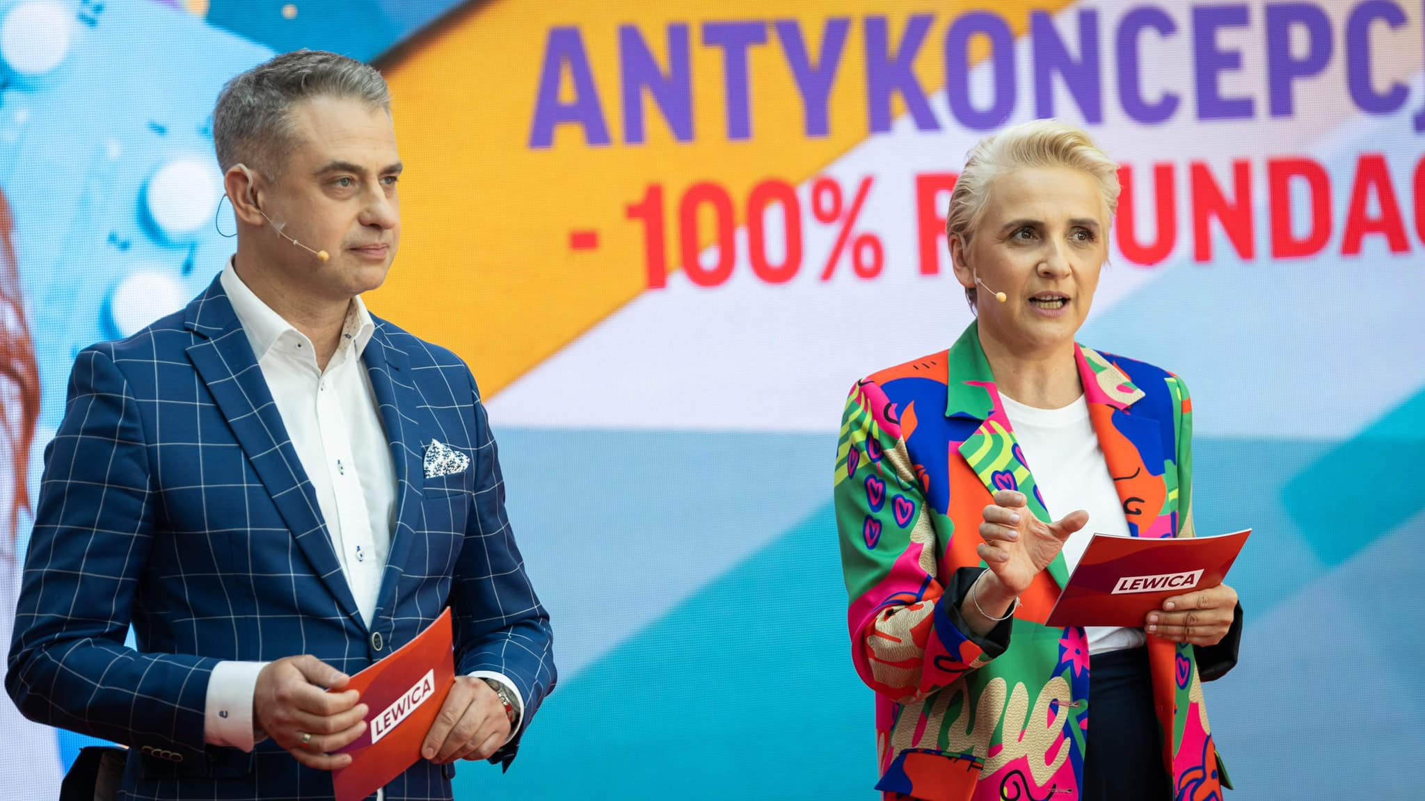 Na scenie dwie osoby prowadzą konwencję „Mamy plan dla mam” Lewicy: mężczyzna w granatowym garniturze w kratę i kobieta w kolorowej marynarce trzymają czerwone kartki z napisem Lewica, a w tle widoczny jest duży, barwny baner.