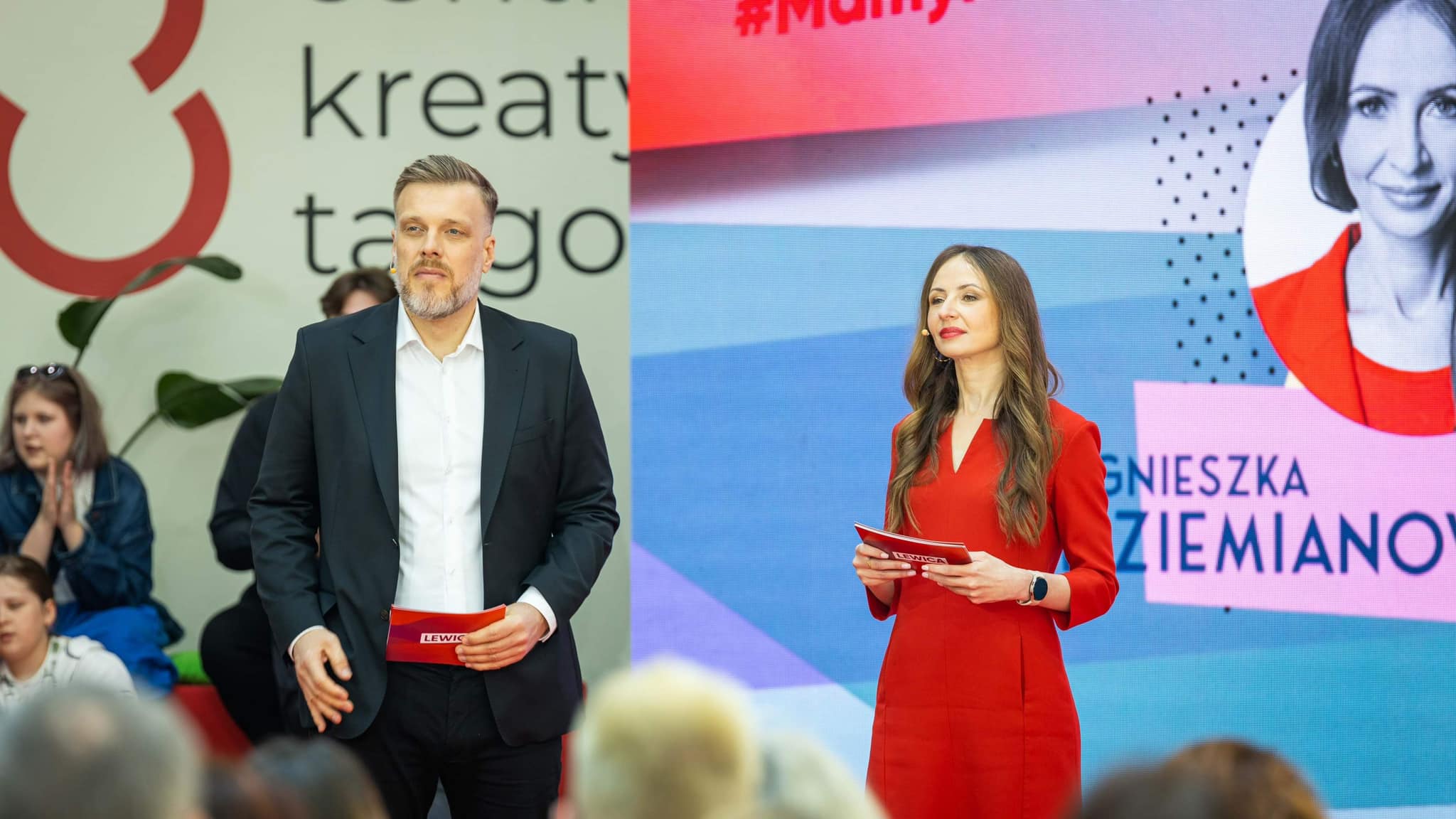 Dwóch prowadzących Konwencji Mamy plan dla mam stoi na scenie: mężczyzna w czarnym garniturze i białej koszuli oraz kobieta w czerwonej sukience trzymają kartki, a za nimi jest kolorowy, duży ekran.