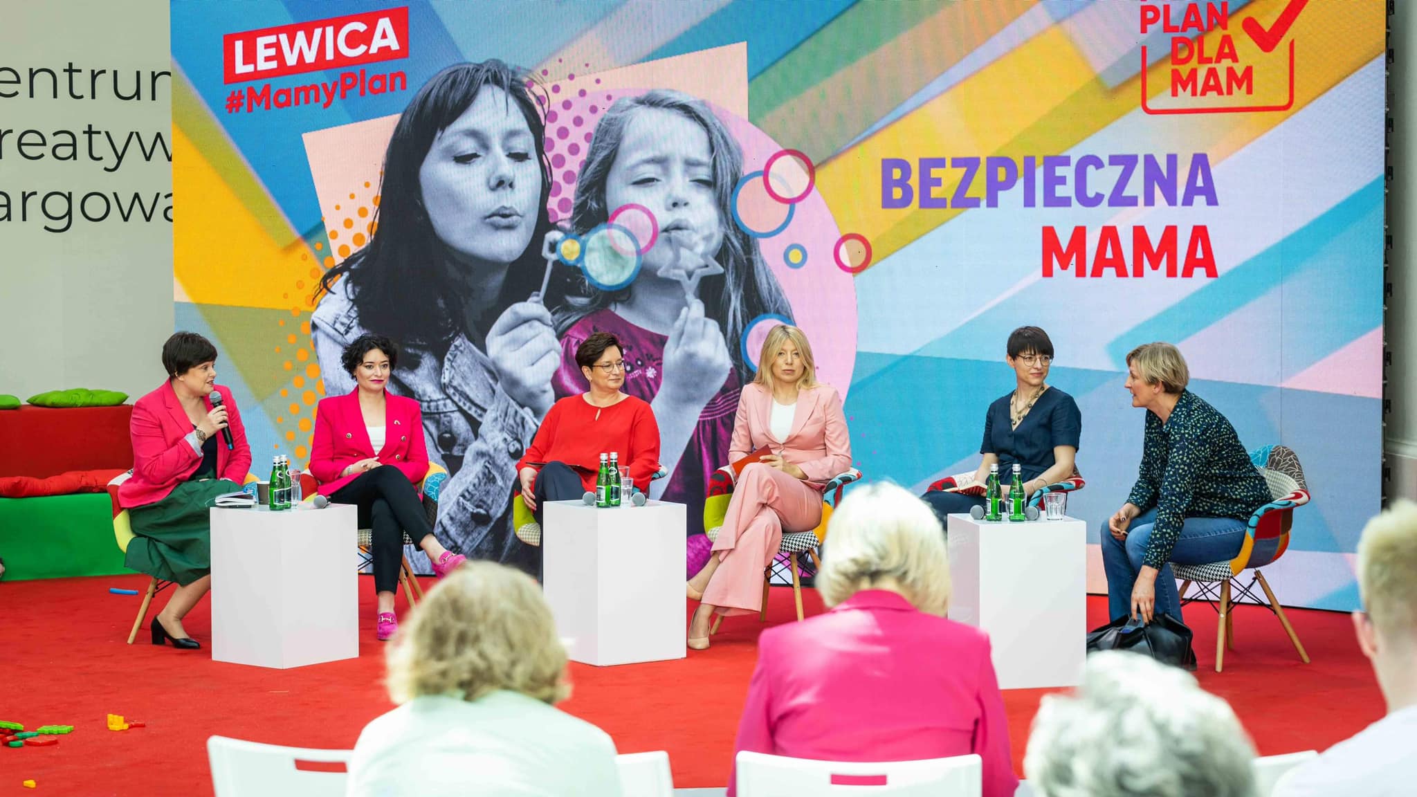Panel dyskusyjny podczas Konwencji Mamy Plan dla Mam, gdzie pięć kobiet siedzi na scenie na czerwonym dywanie przed kolorowym plakatem z hasłami BEZPIECZNA MAMA i #MamyPlan.