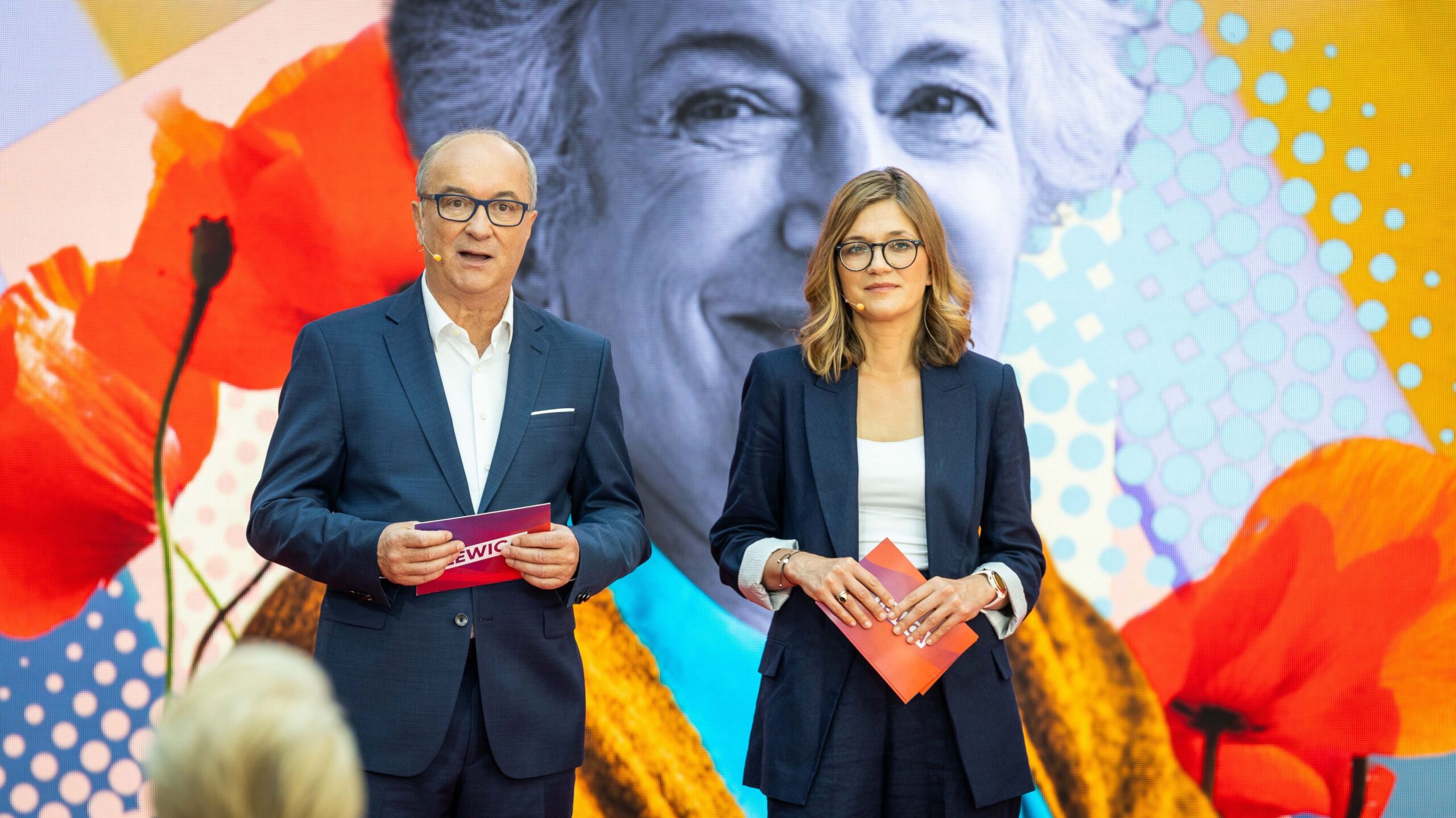 Dwóch prowadzących w granatowych garniturach stoi na scenie podczas Konwencji „Mamy plan dla mam”, trzymając kartki, a w tle widoczna jest kolorowa, abstrakcyjna grafika.