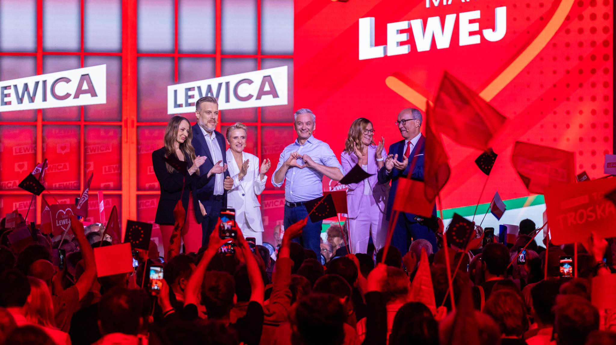 Grupa polityków stoi na scenie podczas wiecu Lewicy, na czerwonym tle z logo Lewica, a centralny mężczyzna formuje serce dłoniami po lewej stronie sceny, podczas gdy tłum z flagami i telefonami wiwatuje.