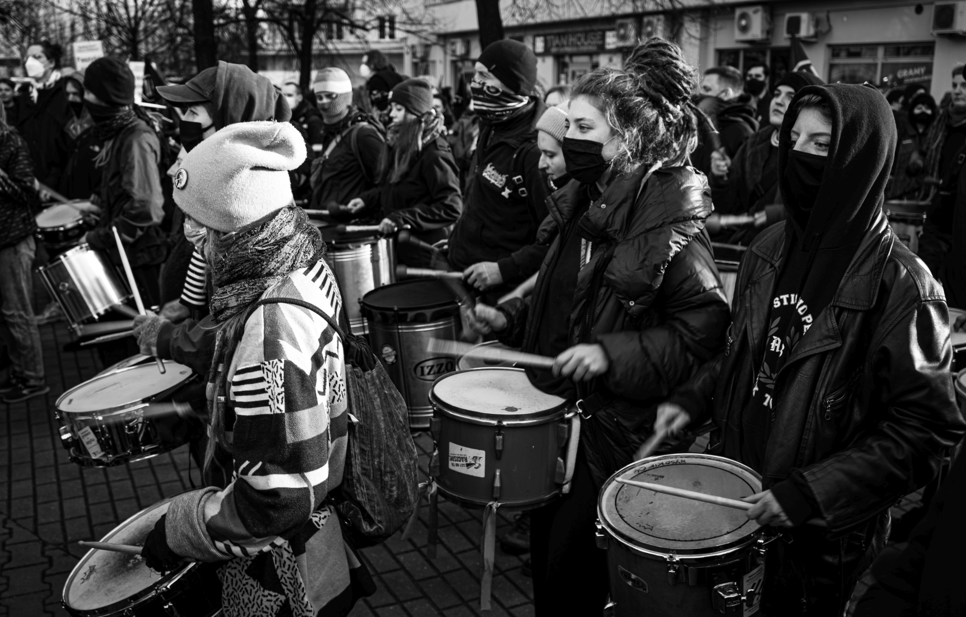 Na czarno-białym zdjęciu grupa ludzi w czapkach i maskach ustnych gra na bębnach podczas marszu.