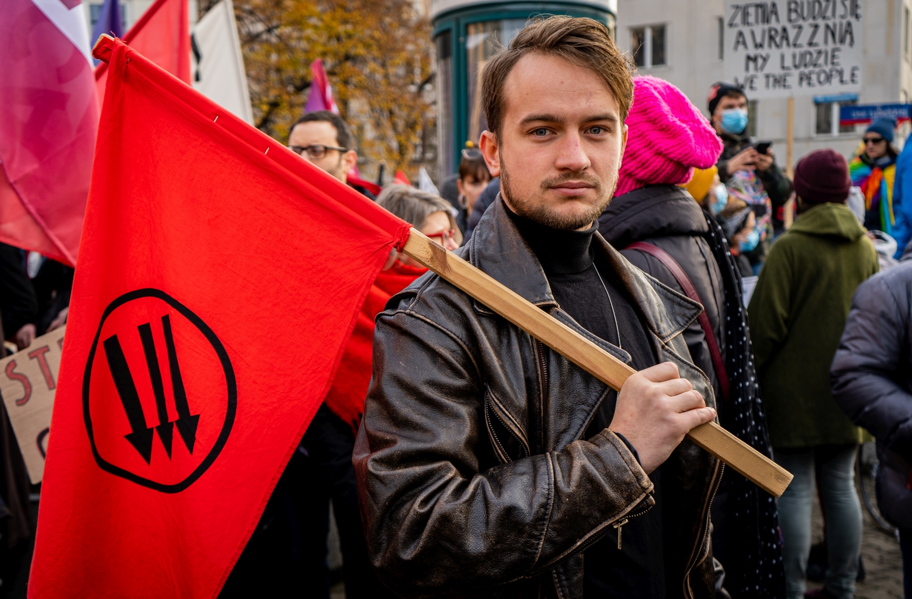 Młody mężczyzna trzyma czerwoną flagę z symbolem trzech strzałek podczas demonstracji.