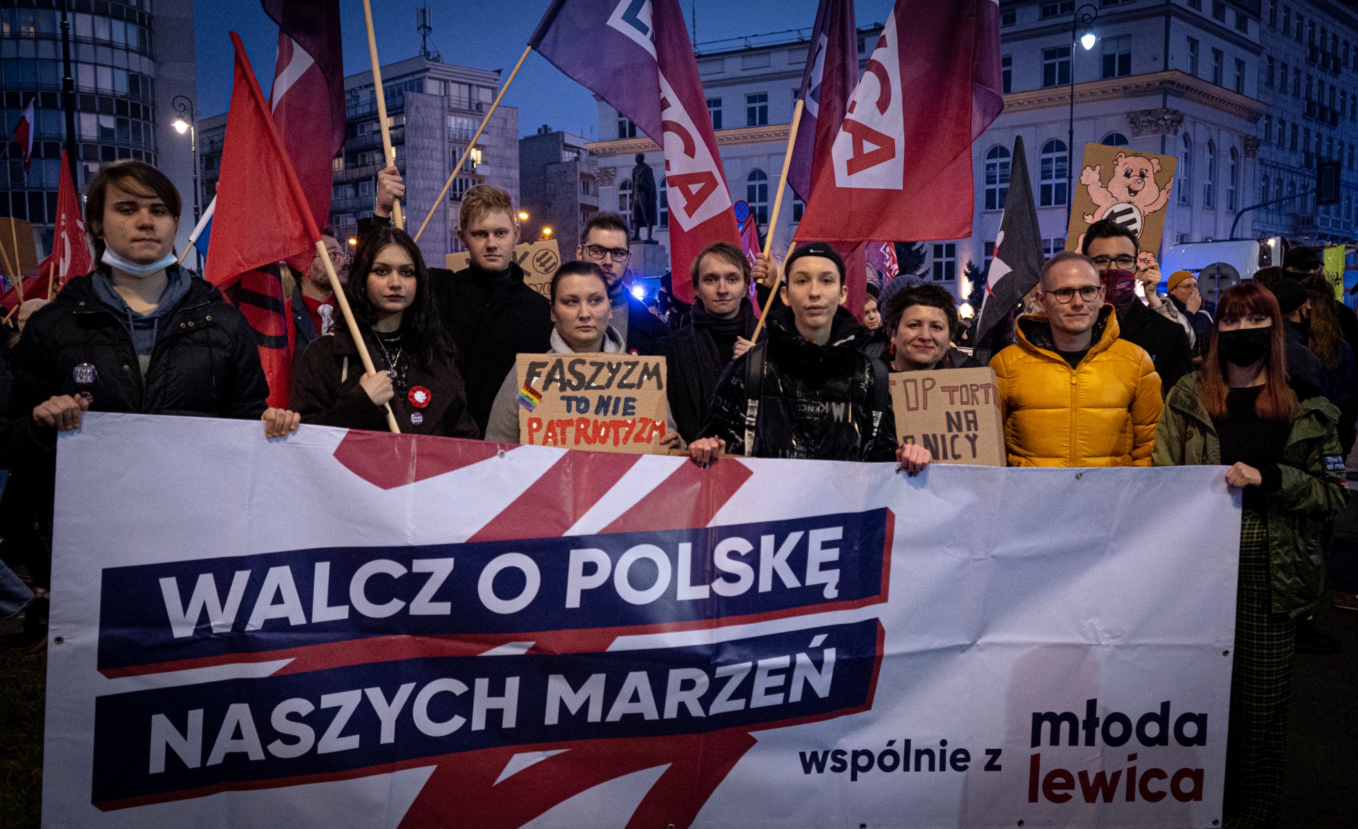 Grupa młodych ludzi uczestniczy w demonstracji trzymając transparenty i flagi, wyrażając swoje postulaty społeczne i polityczne.