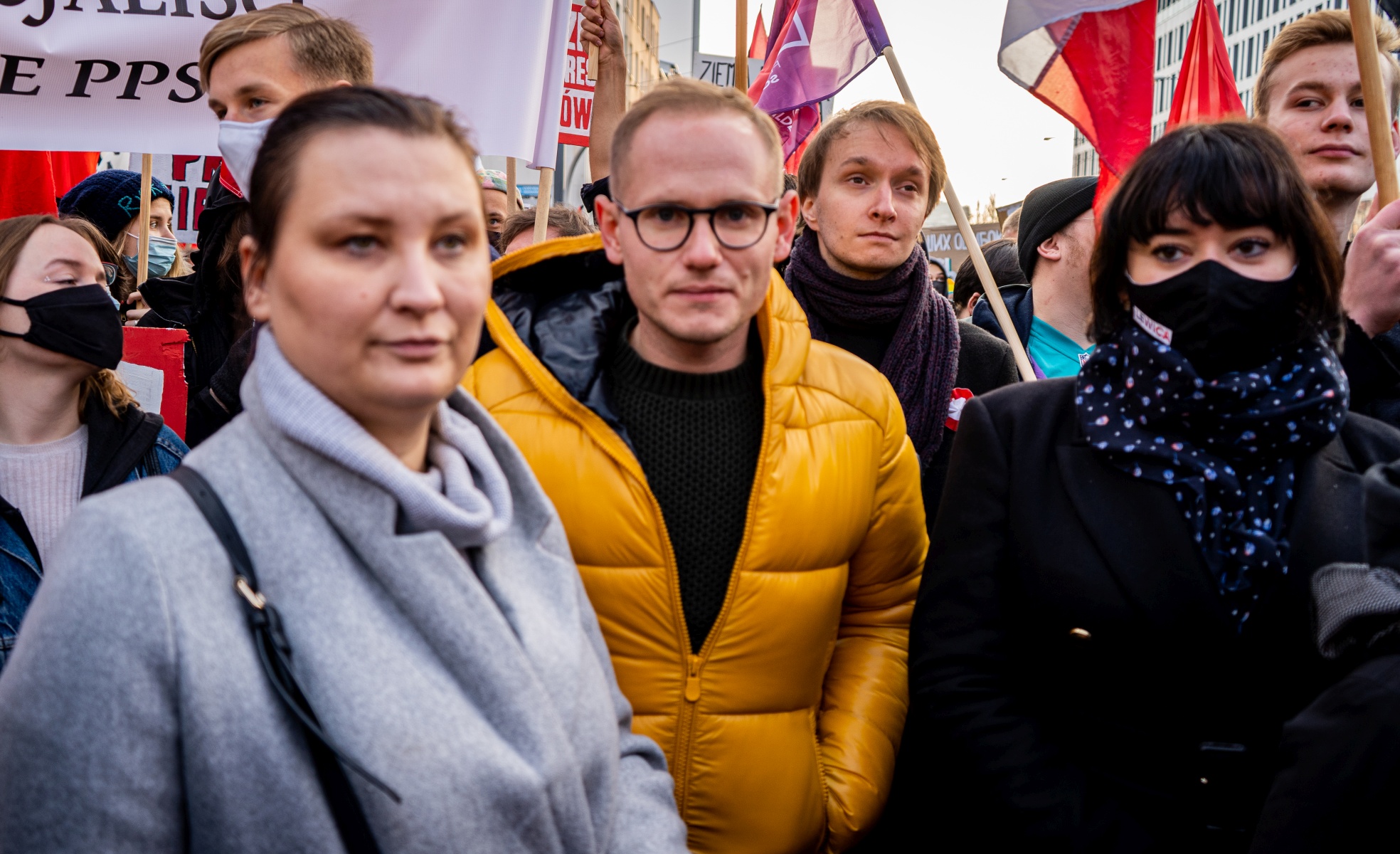 Grupa ludzi uczestniczy w demonstracji, wyrażając swoje postulaty, podczas gdy jedna z osób w żółtej kurtce znajduje się na pierwszym planie.