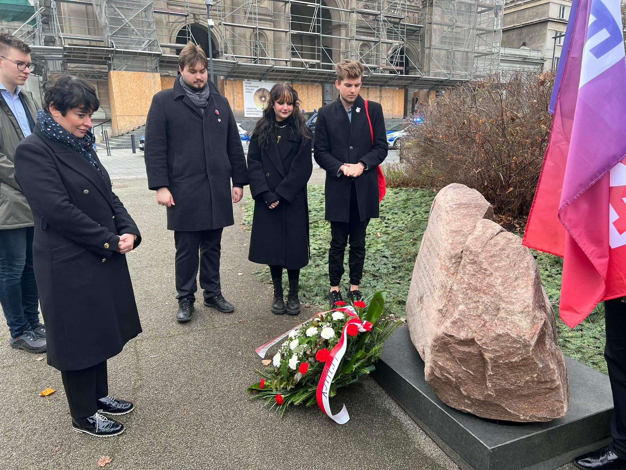 Na zdjęciu grupa osób w czarnych płaszczach składa wieniec przy pomniku, w tle widoczne są flagi i prace budowlane.