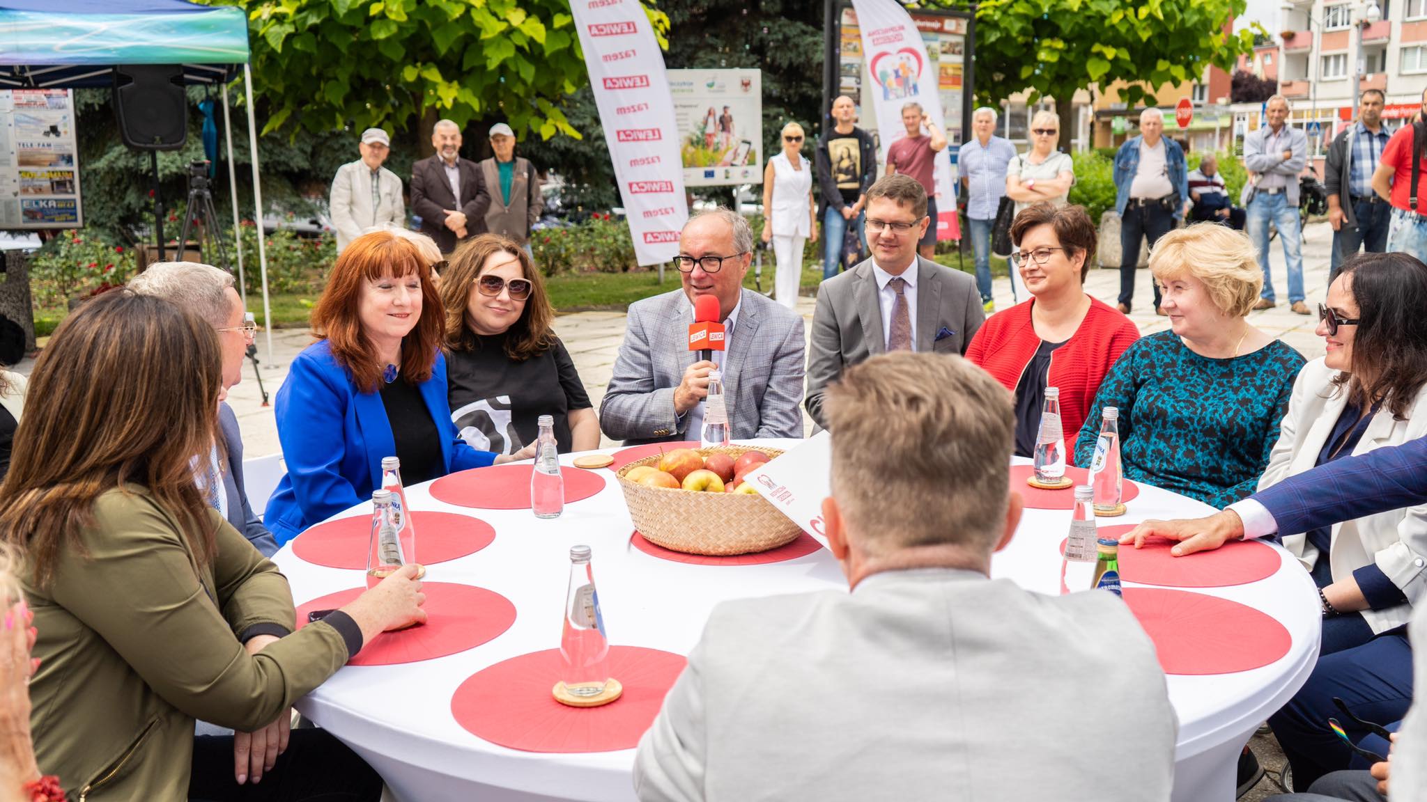 Zdjęcie przedstawia grupę osób siedzących przy okrągłym stole, w trakcie spotkania na świeżym powietrzu, z wyraźnym zainteresowaniem na ich twarzach, w otoczeniu zieleni i z widownią w tle.