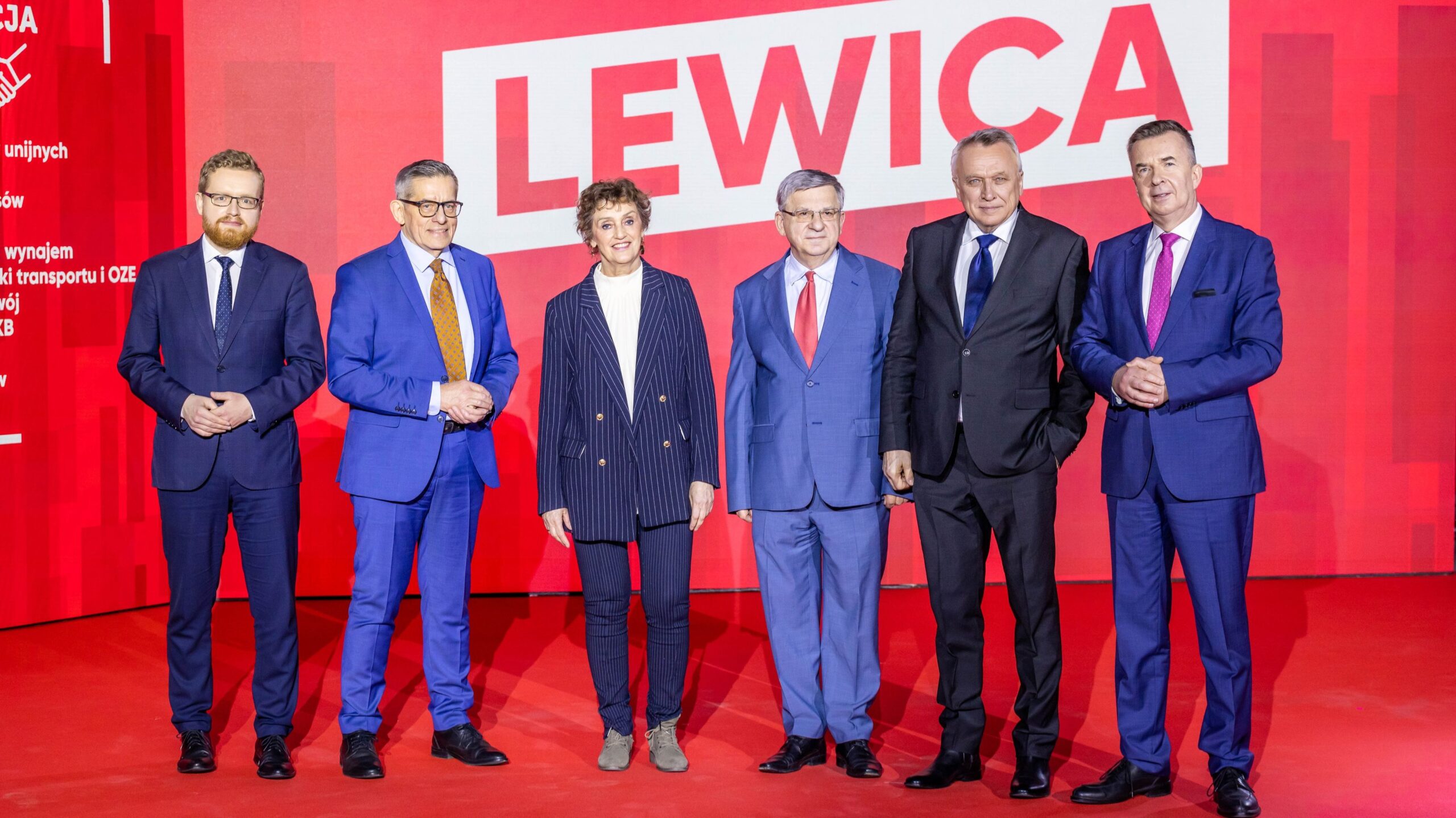 Na zdjęciu widocznych jest sześciu mężczyzn i jedna kobieta stojących obok siebie na scenie, w tle z czerwonym napisem 