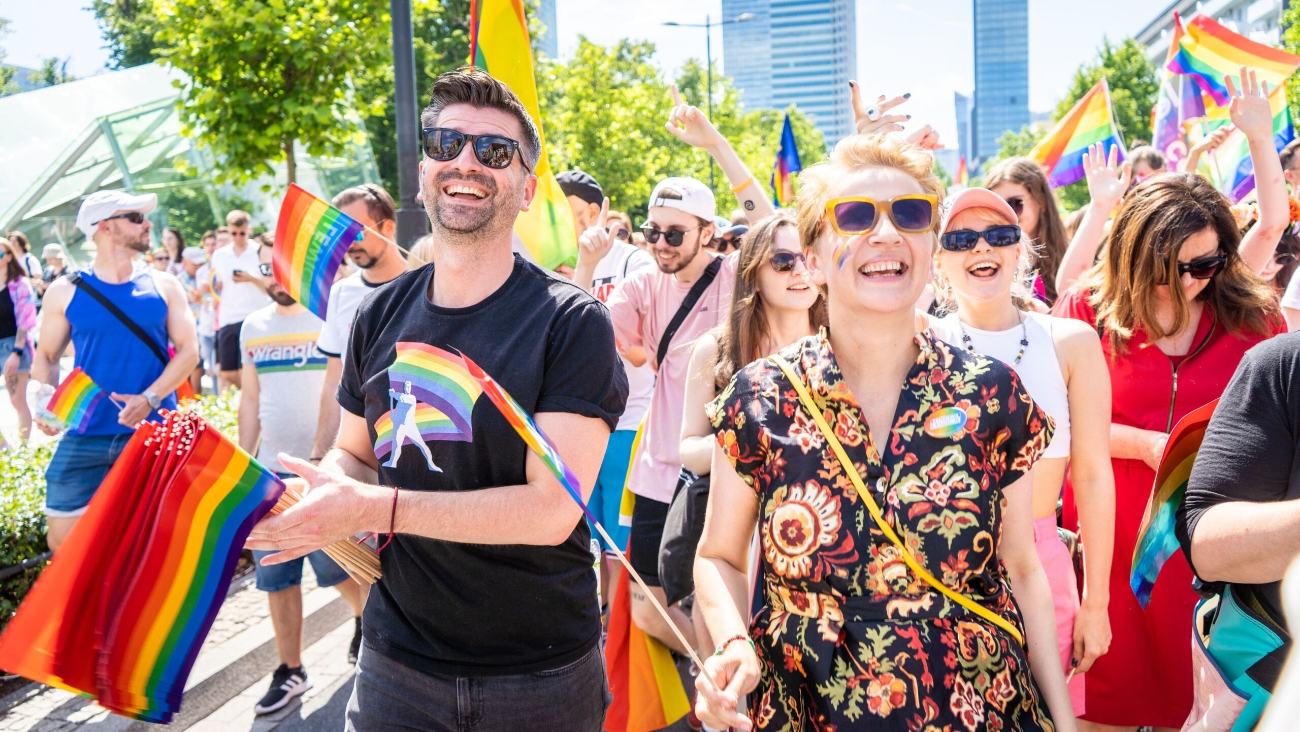 Tłum ludzi w radosnym pochodzie niosącym flagi w kolorach tęczy, celebrujących różnorodność i równość.