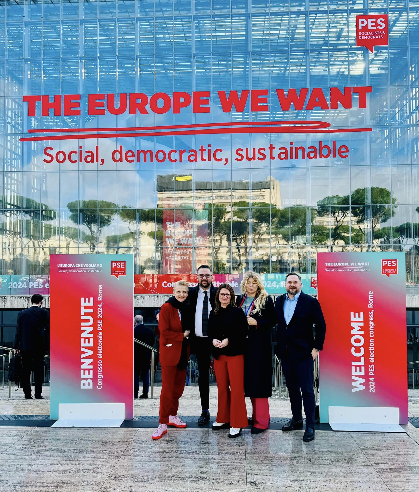 Pięcioosobowa grupa osób w eleganckich ubraniach stoi przed szklaną fasadą budynku z dużym czerwonym napisem "THE EUROPE WE WANT" oraz banerami PES.