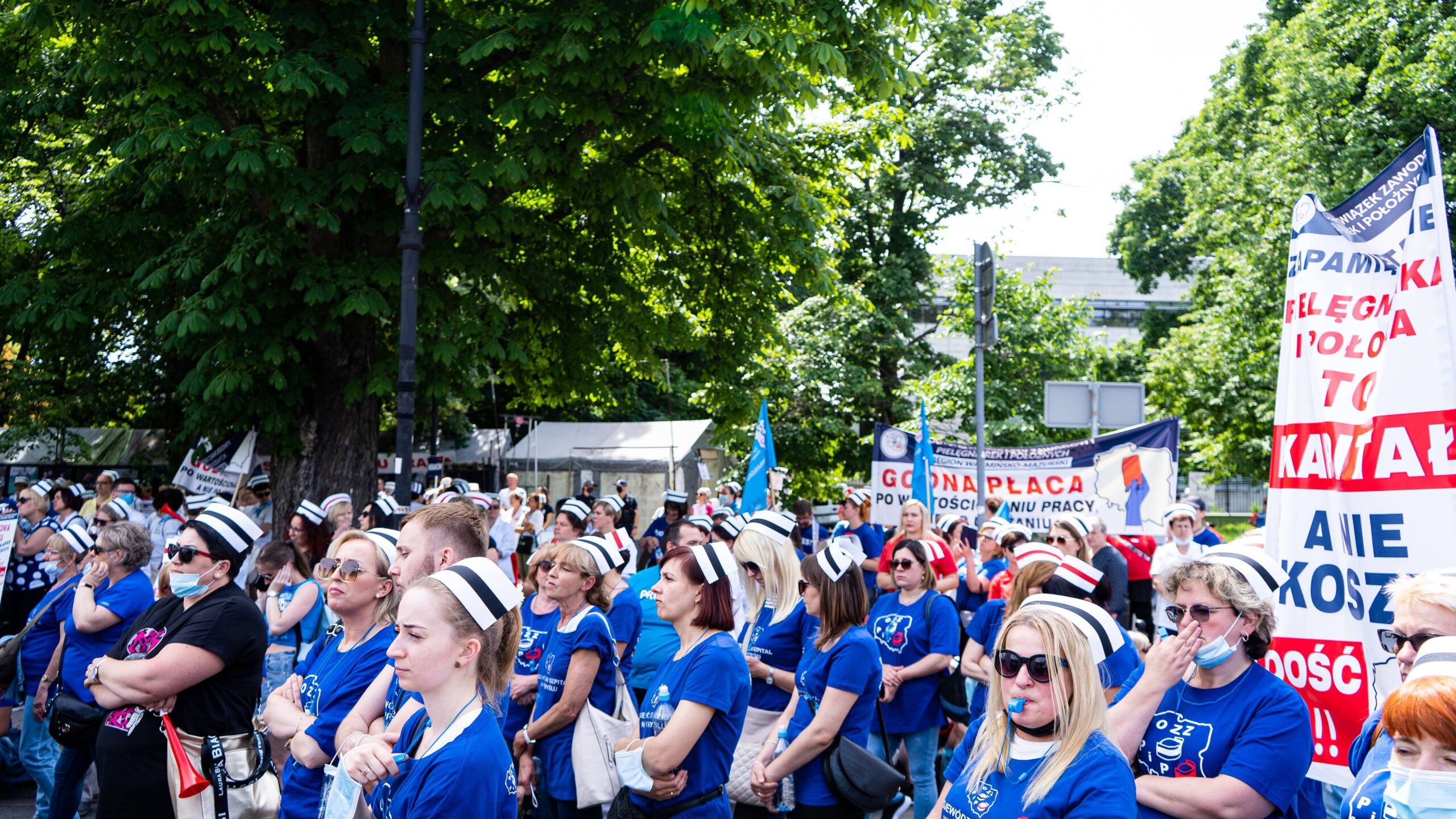 Duża grupa pielęgniarek i położnych w niebieskich koszulkach i biało-niebieskich czapkach protestuje w parku, trzymając banery i flagi.