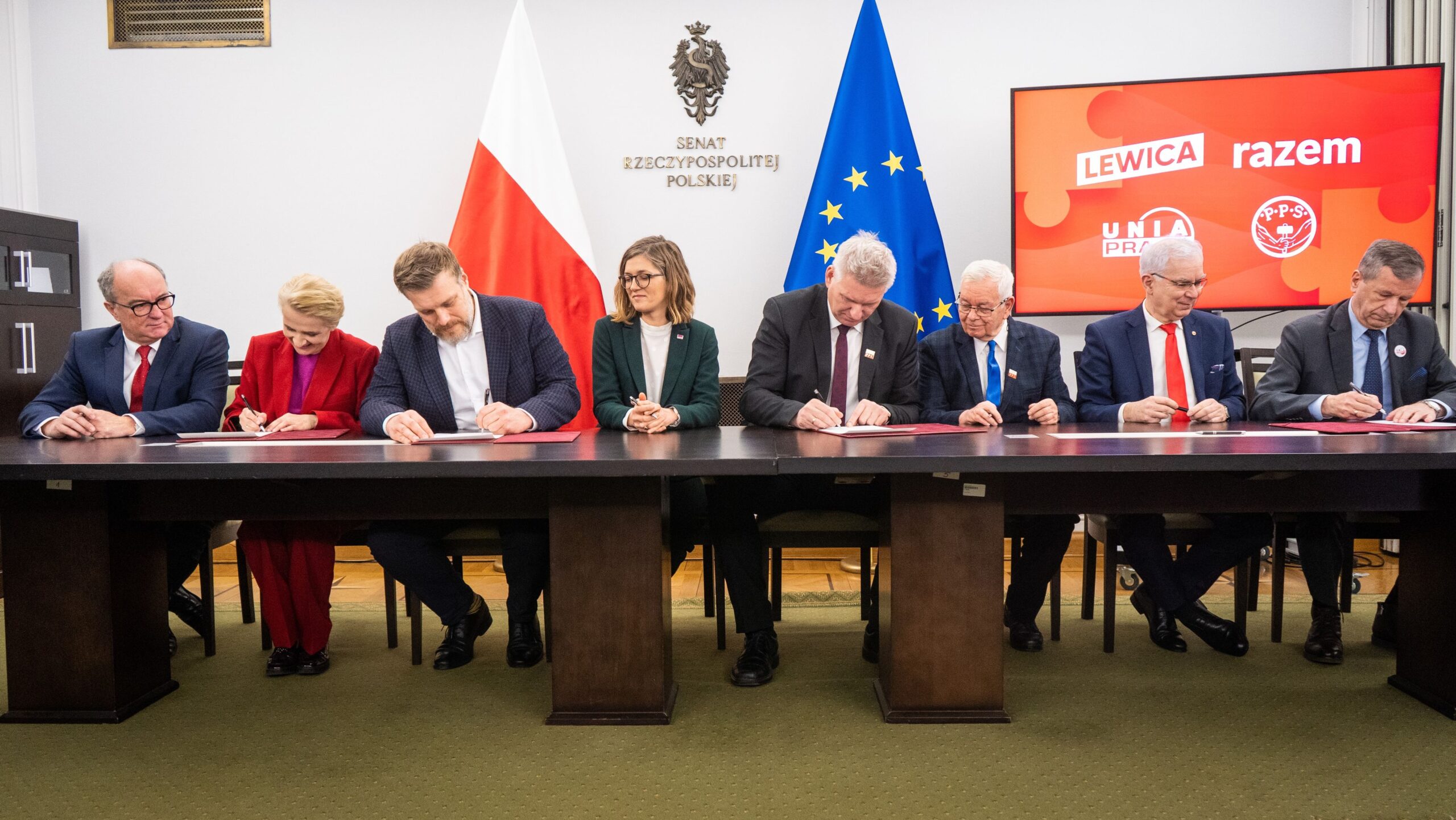 Na zdjęciu widoczni są politycy podpisujący dokumenty w sali Senatu, z flagami Polski i Unii Europejskiej w tle.