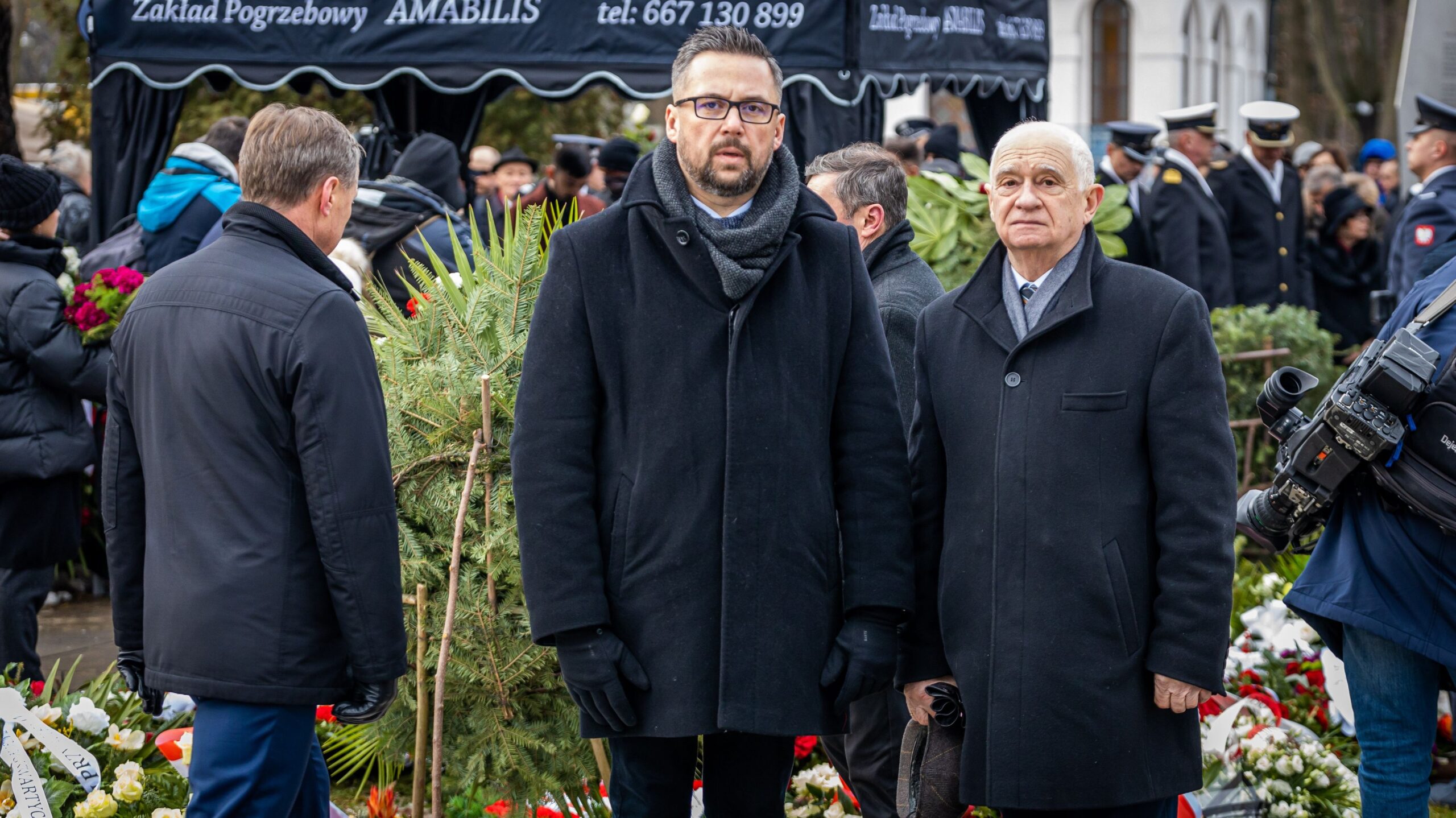 Na zdjęciu dwóch mężczyzn w czarnych płaszczach stoi na pogrzebie, w tle kondukt żałobny, kwiaty i ekipa filmowa.