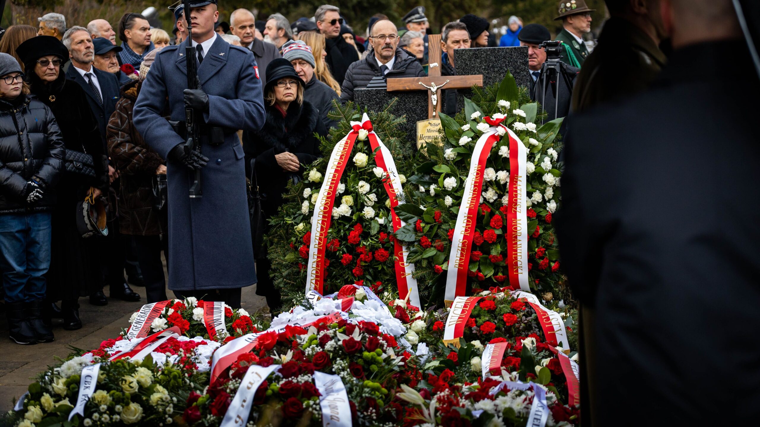 Na zdjęciu widoczny jest pogrzeb, w którym uczestniczy wielu ludzi, a przed nimi znajdują się kwiaty oraz krzyż.