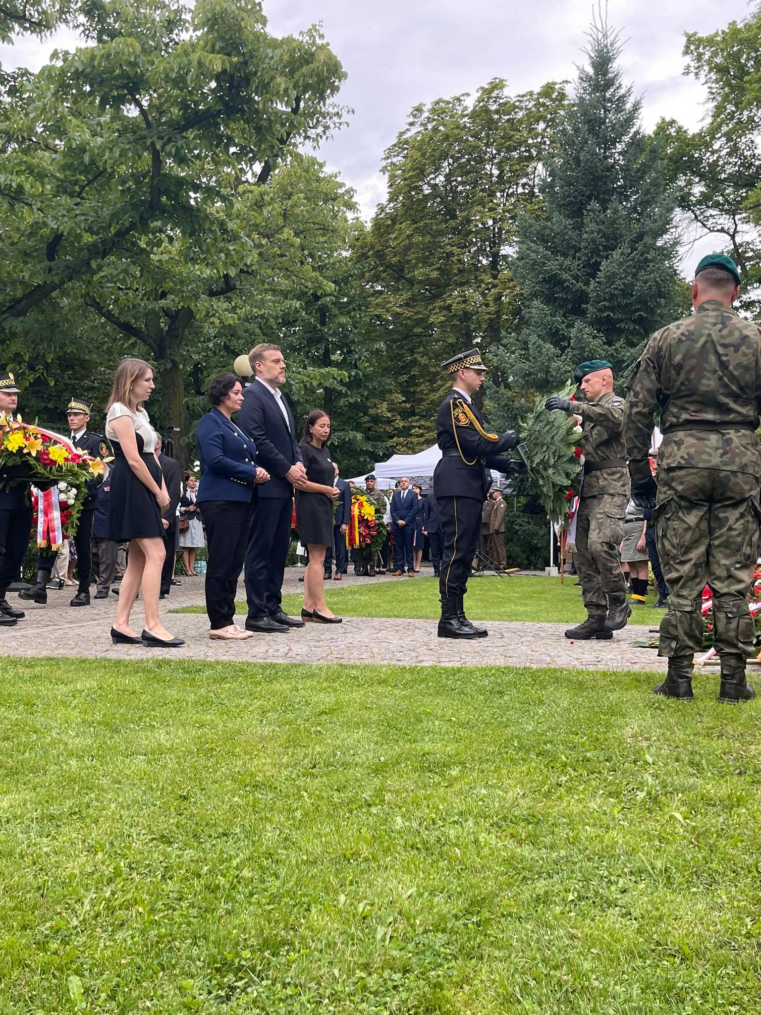 Uczestnicy uroczystości w parku składają hołd bohaterom Powstania Warszawskiego — żołnierze w mundurach i cywile w eleganckich ubraniach stoją przy wiązance kwiatów.