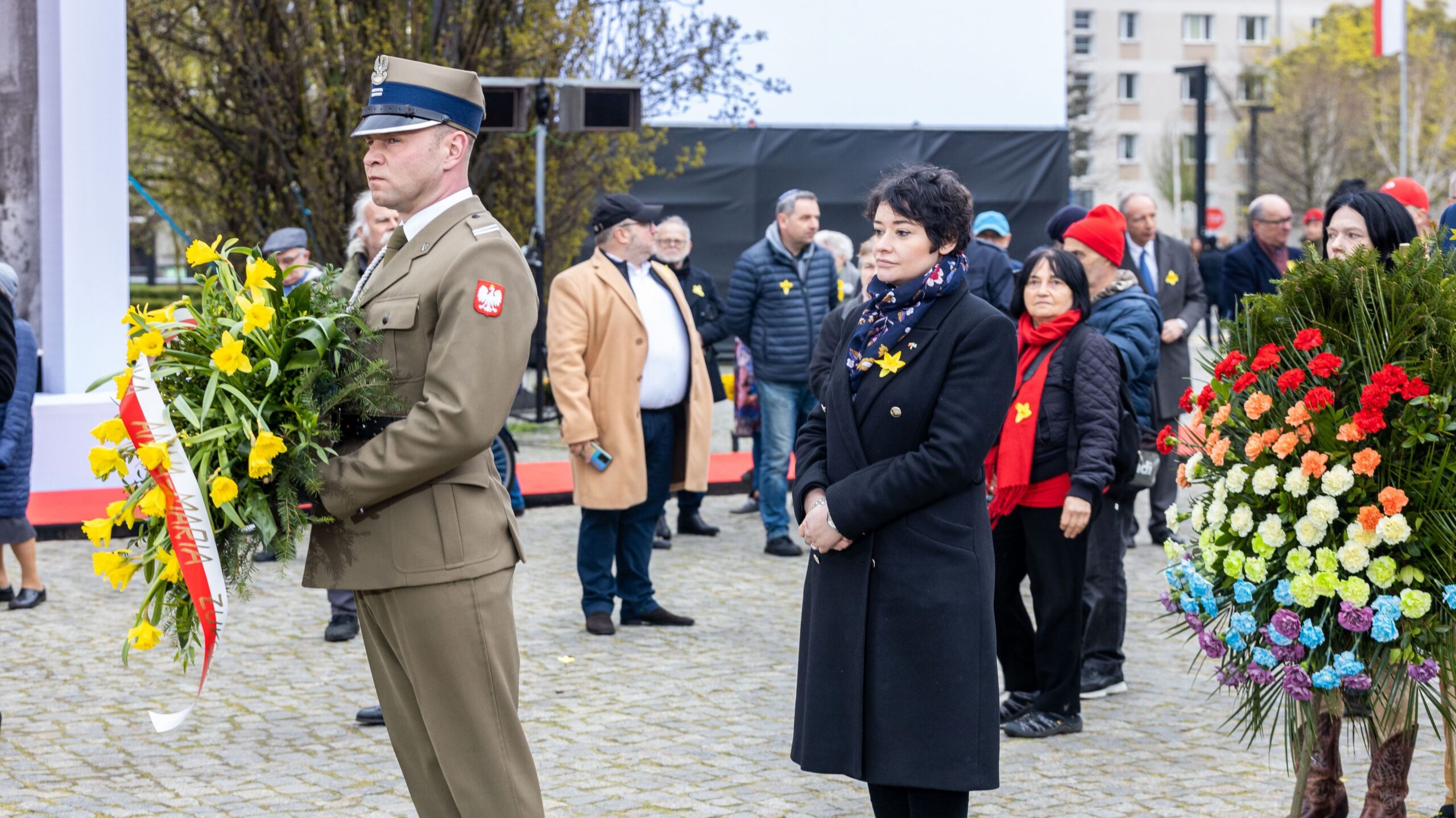 Na zdjęciu widoczny jest żołnierz w mundurze z wiązanką kwiatów oraz grupa osób biorących udział w uroczystości, z kwiatami w rękach, na tle miejskiego krajobrazu.