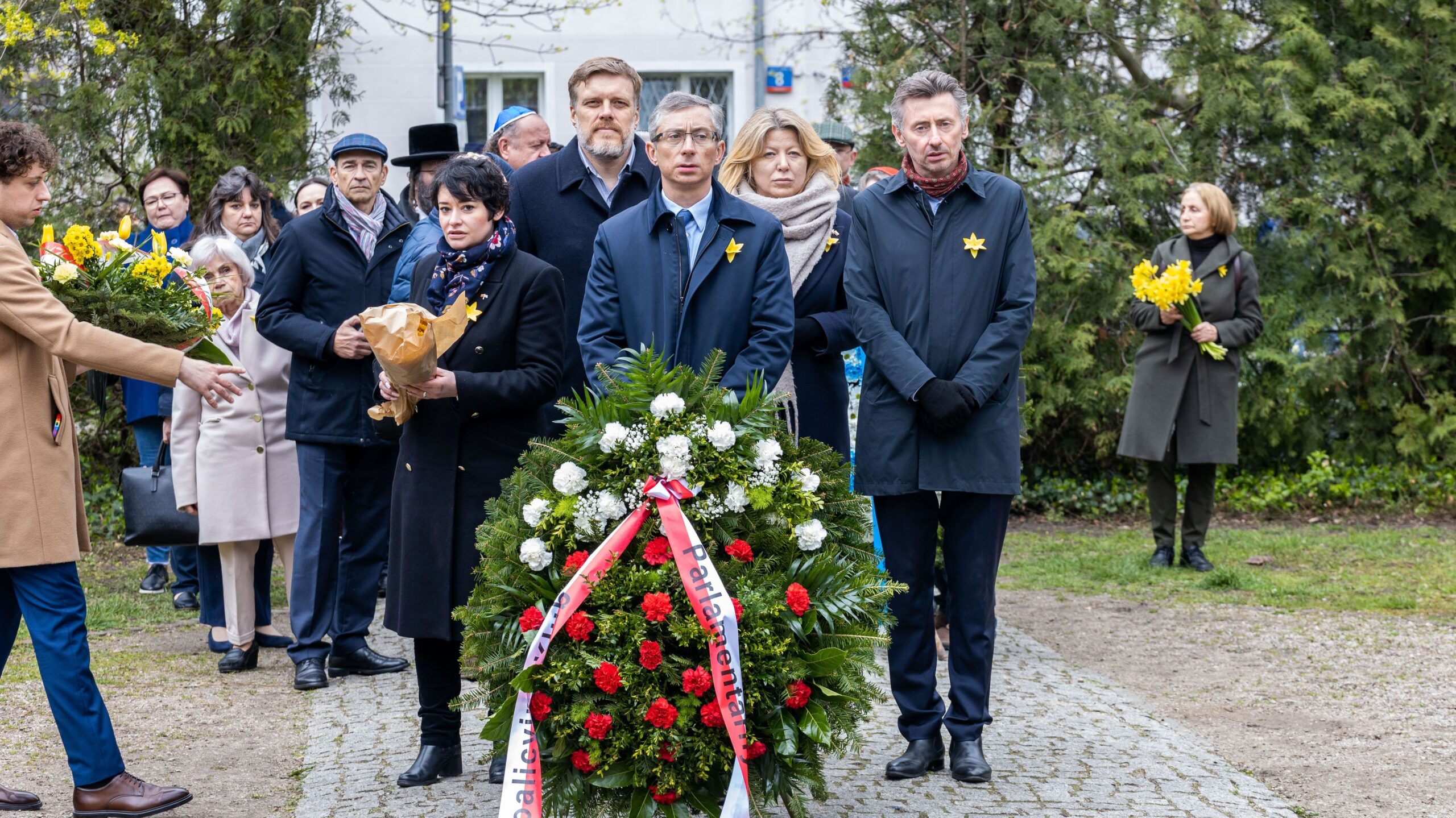 Na zdjęciu widoczna jest grupka ludzi zgromadzonych wokół wieńca kwiatowego, uczestniczących w uroczystości upamiętniającej.