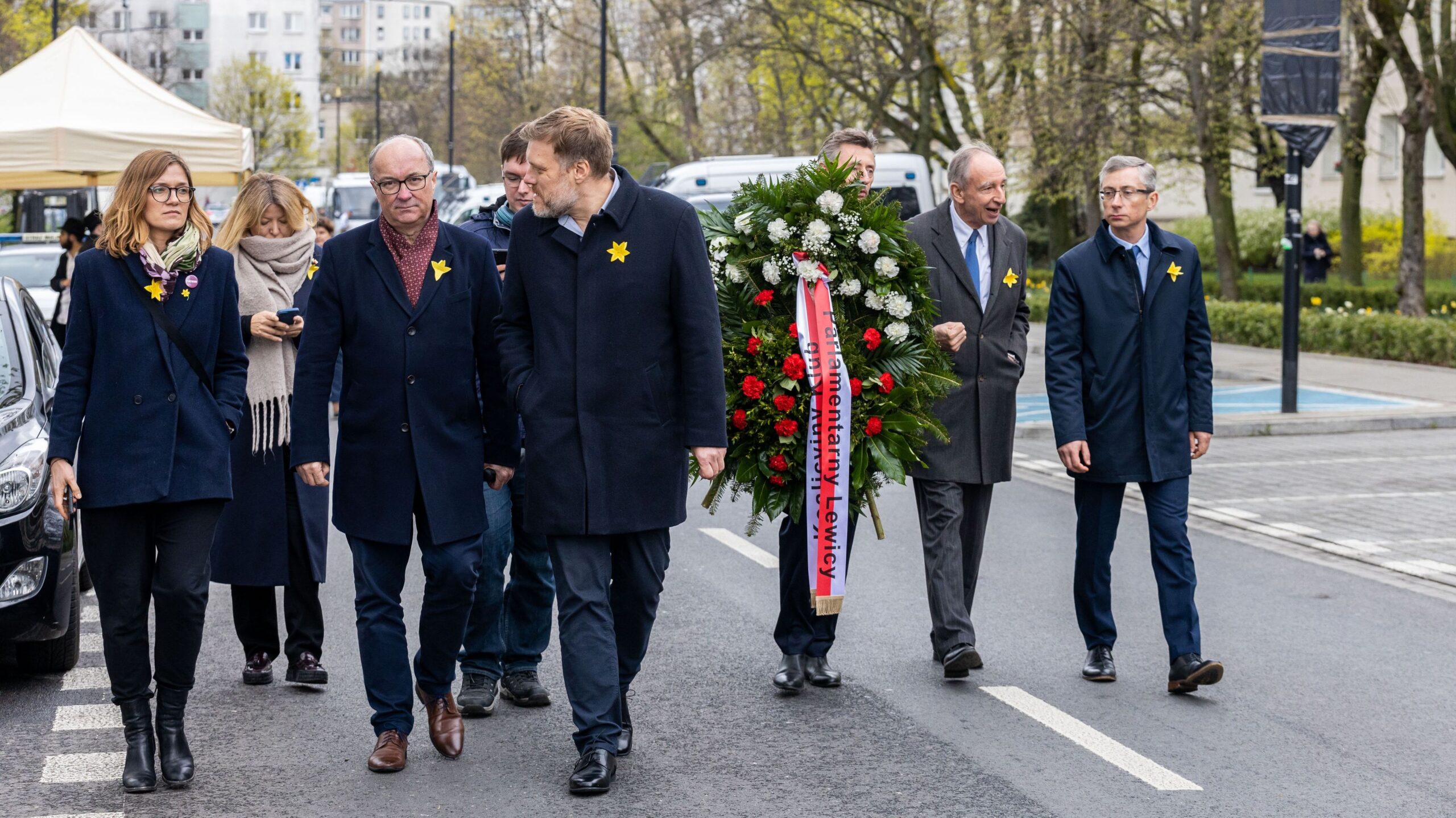 Grupa osób w garniturach z żółtymi miniaturowymi żonkilami na piersiach idzie w kierunku kamery, niosąc wieniec z czerwonymi i białymi kwiatami.