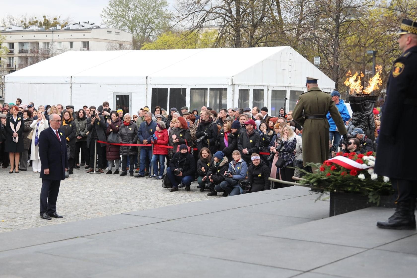 Uczestnicy obchodów 79. rocznicy Powstania w Getcie Warszawskim zgromadzeni na placu, wśród fotografów, przed paleniskiem z ogniem i wieńcem kwiatów przy scenie.
