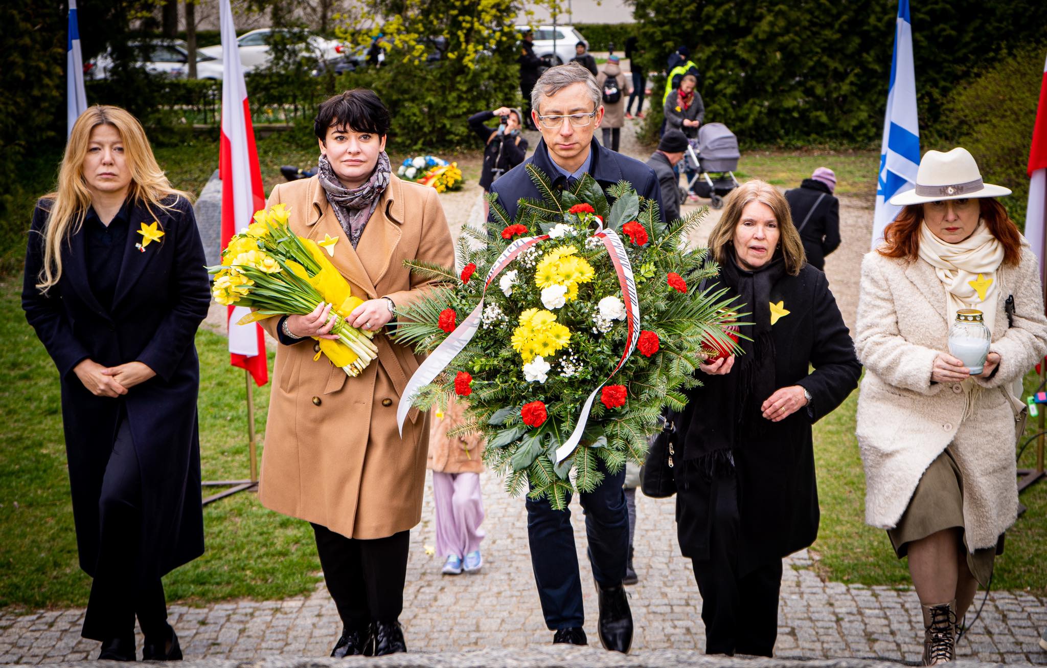 Grupa osób idzie placem, niesie duży wieniec kwiatowy oraz bukiety podczas obchodów 79. rocznicy Powstania w Getcie Warszawskim, z flagami widocznymi w tle.