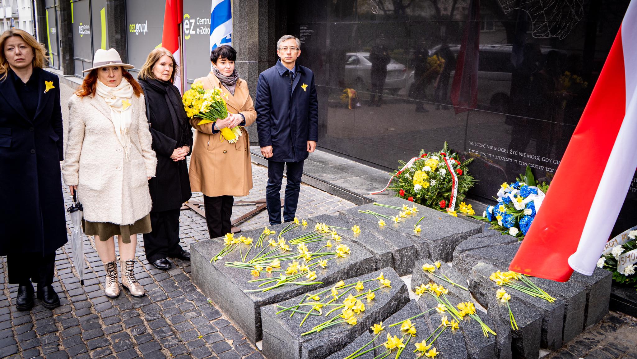 Grupa osób składa żółte żonkile przed pamiątkową ścianą Powstania w Getcie Warszawskim, przy tle flagi Polski.