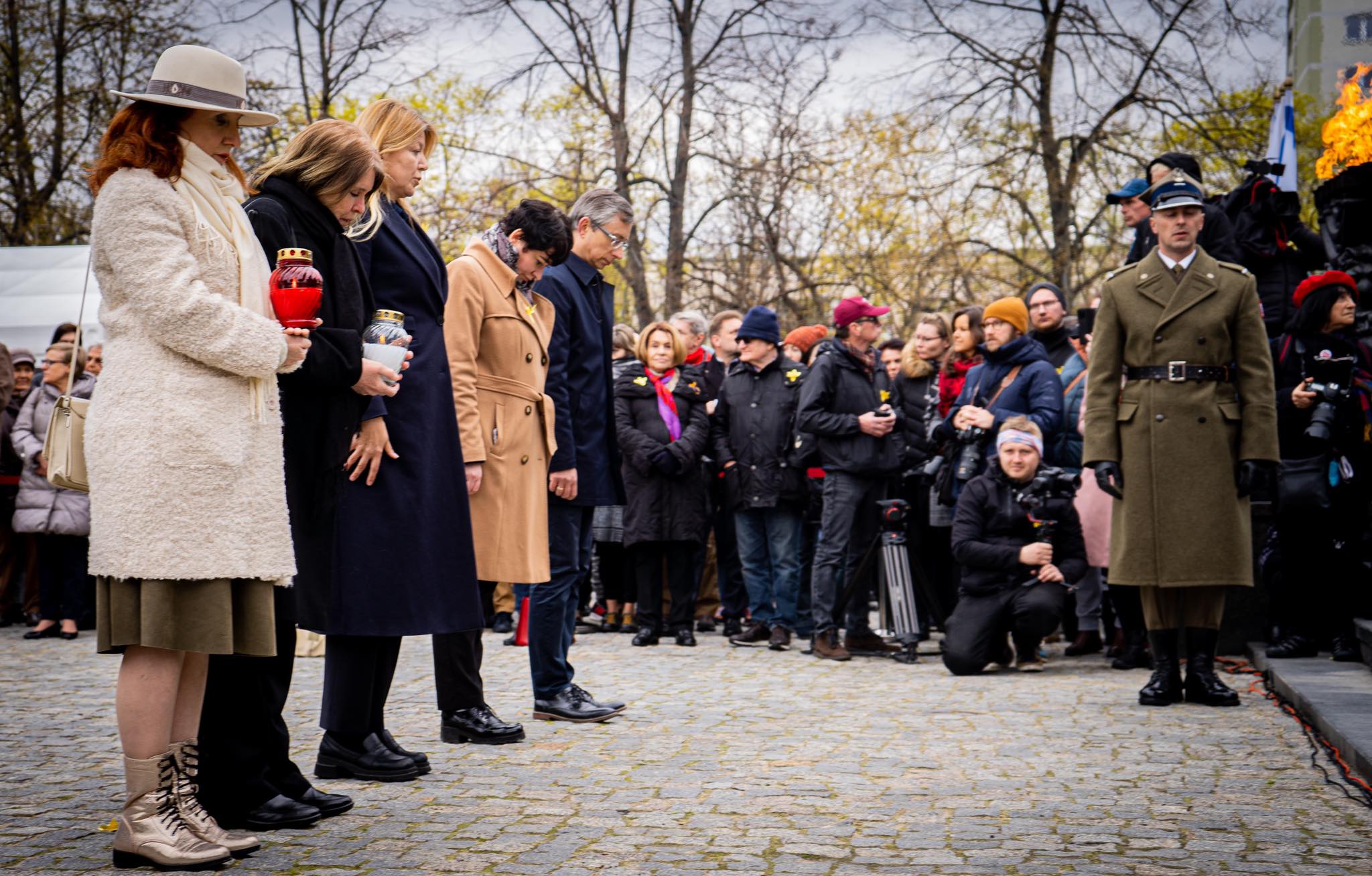 Uczestnicy obchodów 79. rocznicy Powstania w Getcie Warszawskim składają hołd, trzymając zapalone znicze, podczas gdy obok stoi oficer w mundurze, a tłum fotografuje wydarzenie.